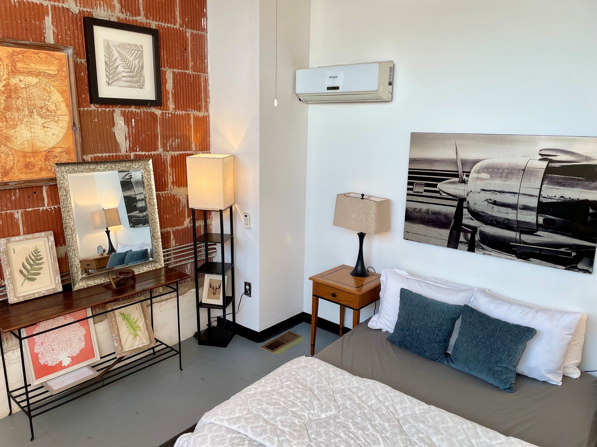 A cozy bedroom corner with a red brick wall decorated with framed botanical art, a large mirror, and a small wooden table with a white table lamp. The room features a white air conditioning unit, a black and white airplane-themed photograph, and a be