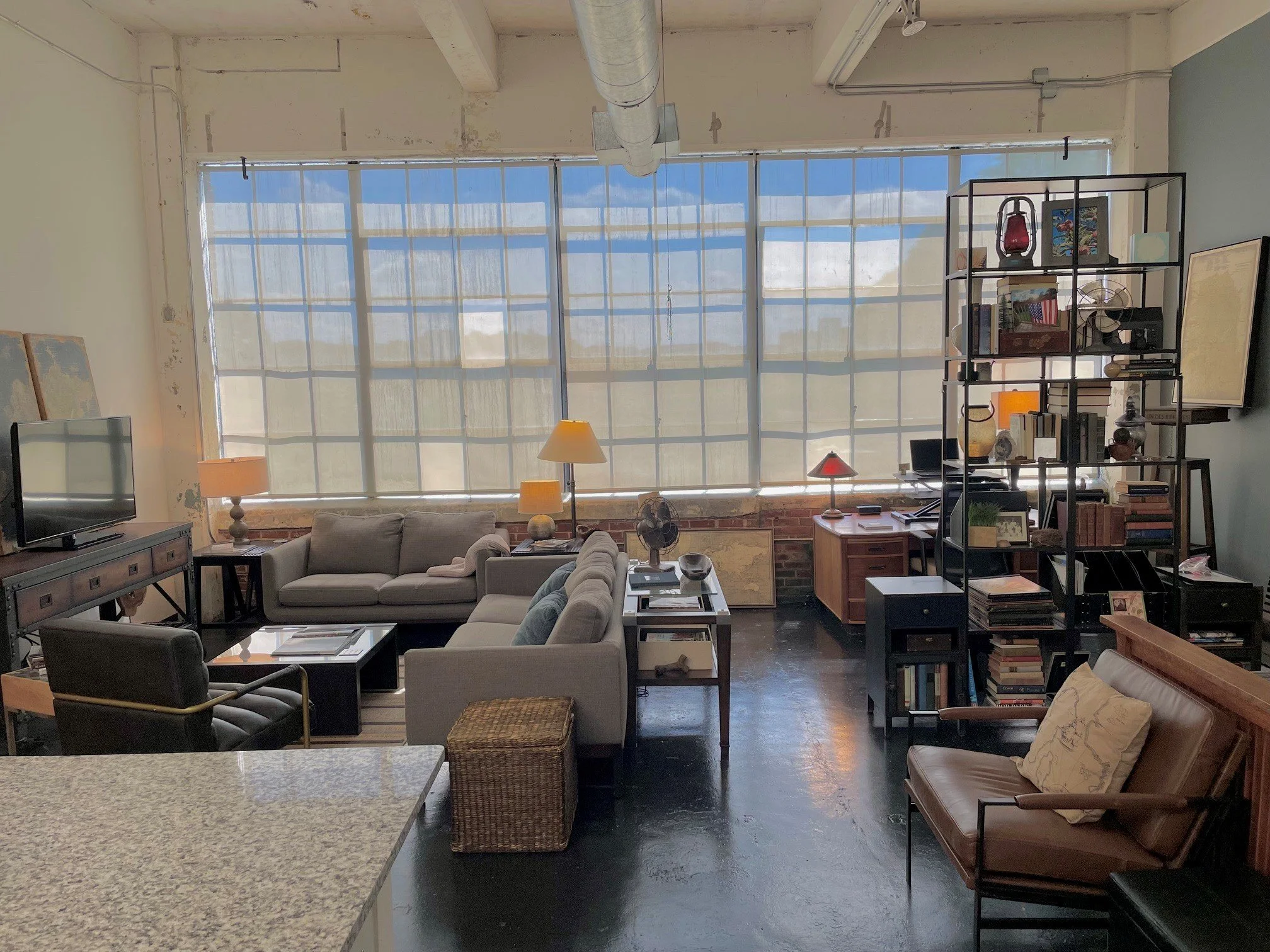 Living room with large window, gray sofas, wooden side tables, a black metal shelving unit filled with books, decor, and a lamp, a small television, and various lamps, with exposed brick and unfinished walls.