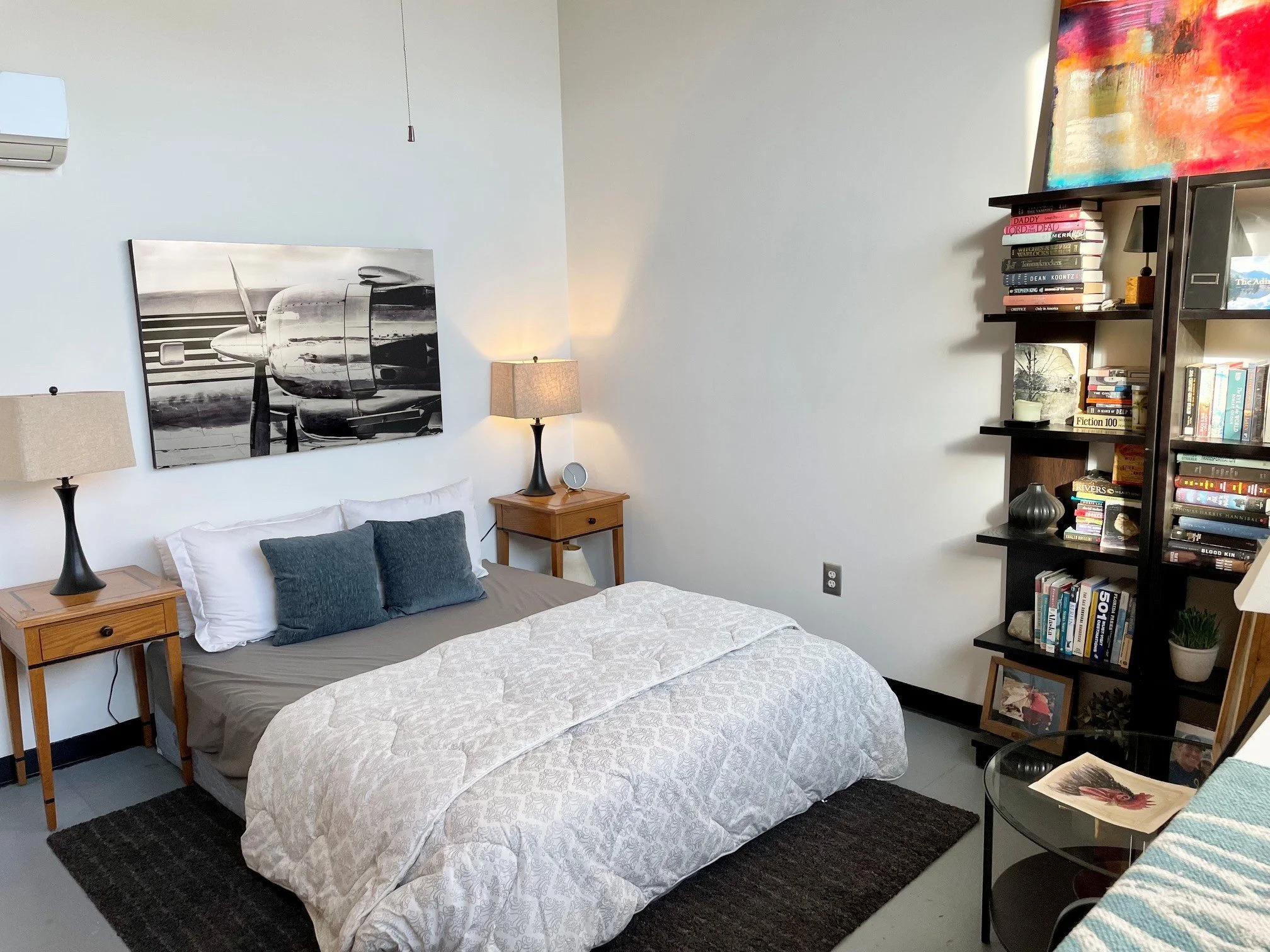 A bedroom with white walls, a bed with a beige patterned quilt and gray and white pillows, two matching bedside tables with lamps, a black bookshelf filled with books and decorative items, a black vase, a framed picture, and a colorful painting on th