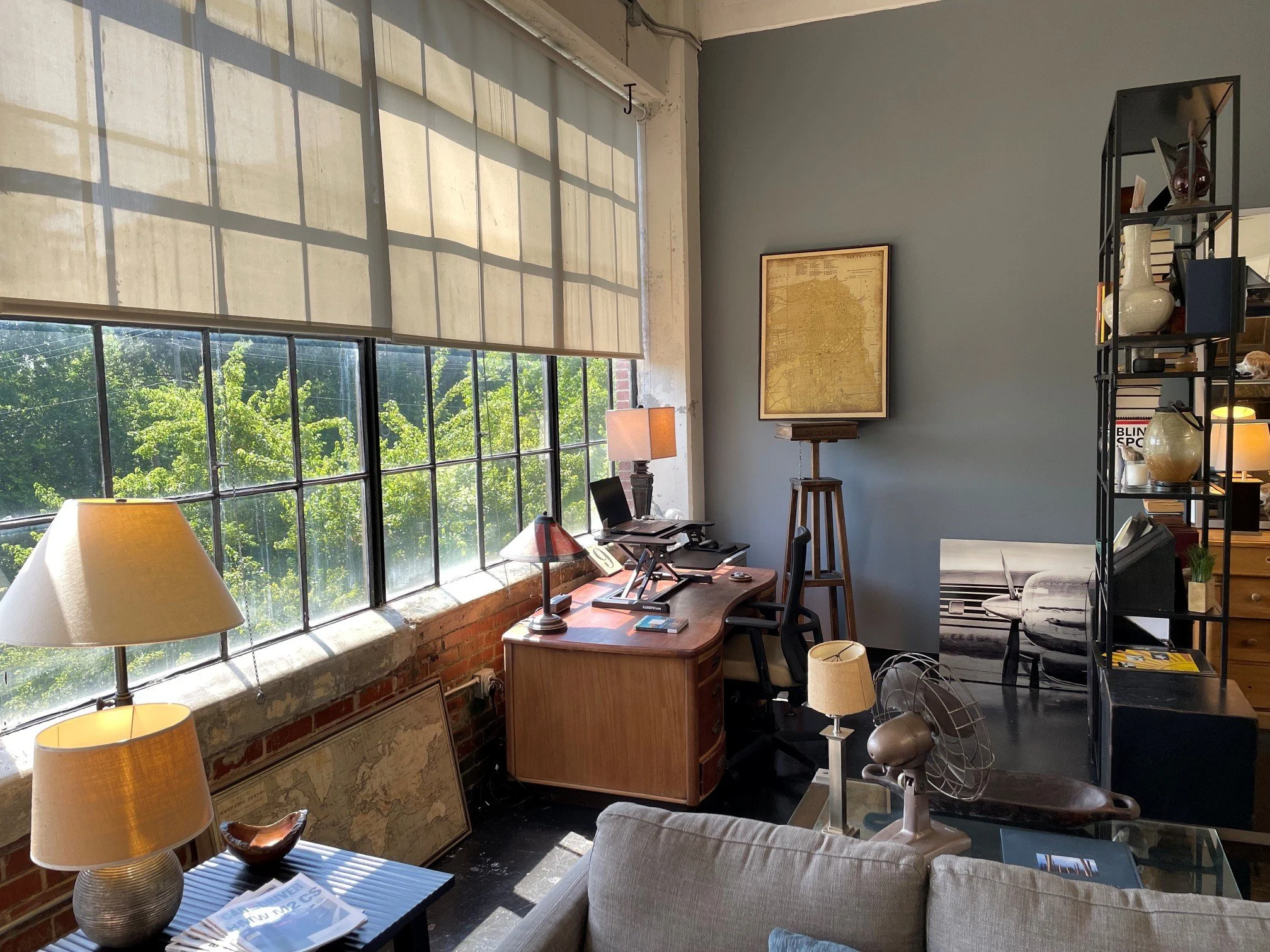 A cozy office corner with large industrial-style windows, a wooden desk, a beige armchair, various lamps, a bookshelf filled with books and decorative items, and a framed map on the wall.