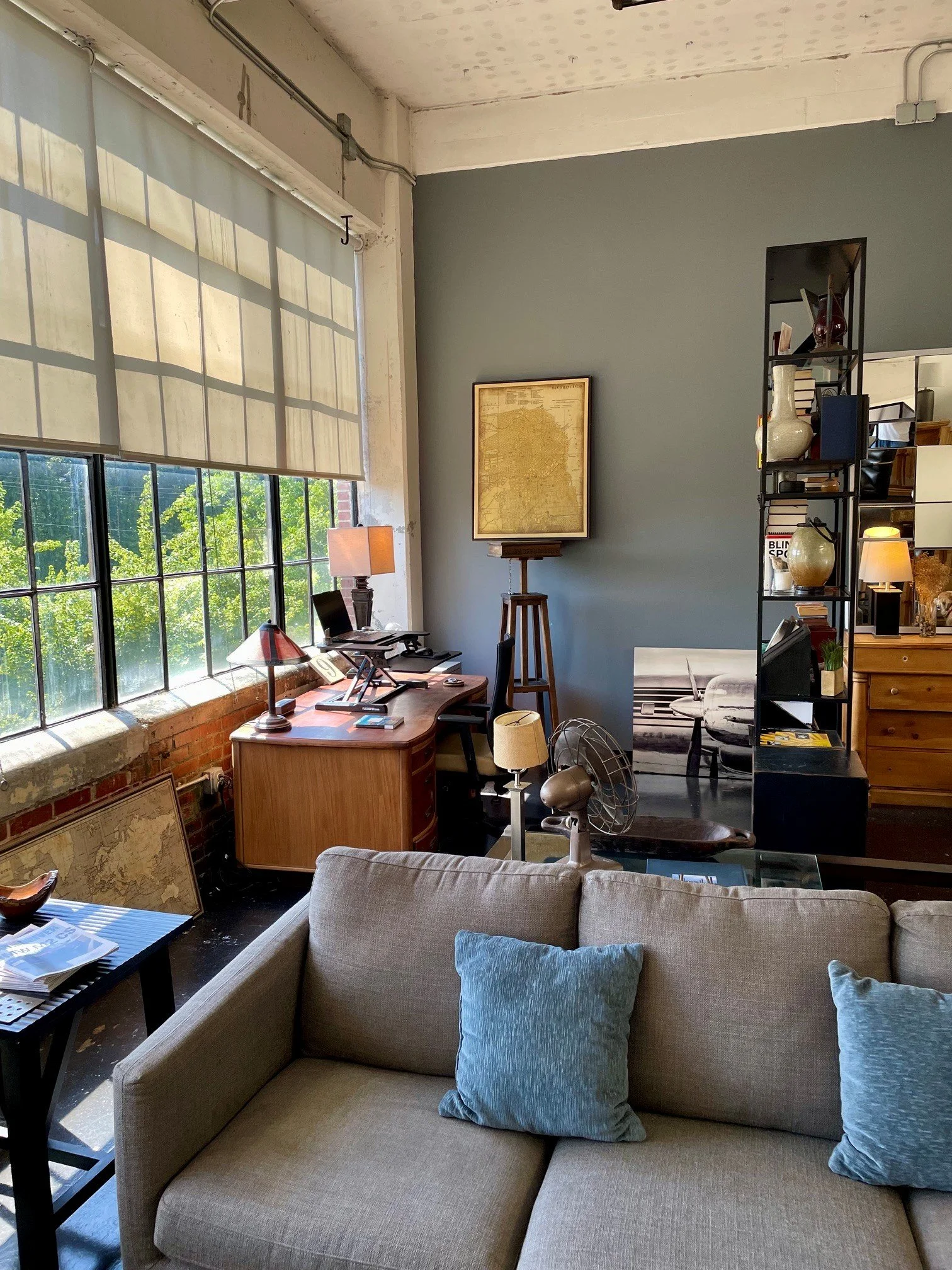 A cozy living room with a beige sofa and blue pillows, a black side table with books and magazines, large windows with blinds allowing sunlight, a wooden desk with a lamp and computer, a wall map, a black metal shelving unit with decorative items, an