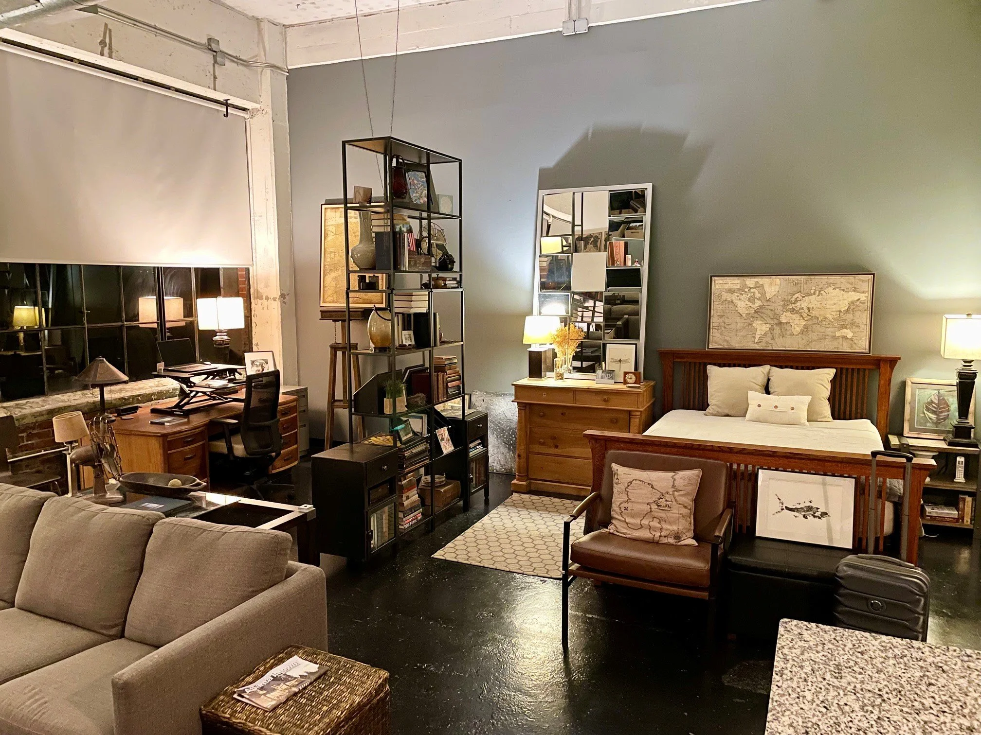 Living room with various furniture including a gray sofa, a leather armchair, a bed, bookshelves, lamps, framed artwork, and a world map on the wall.
