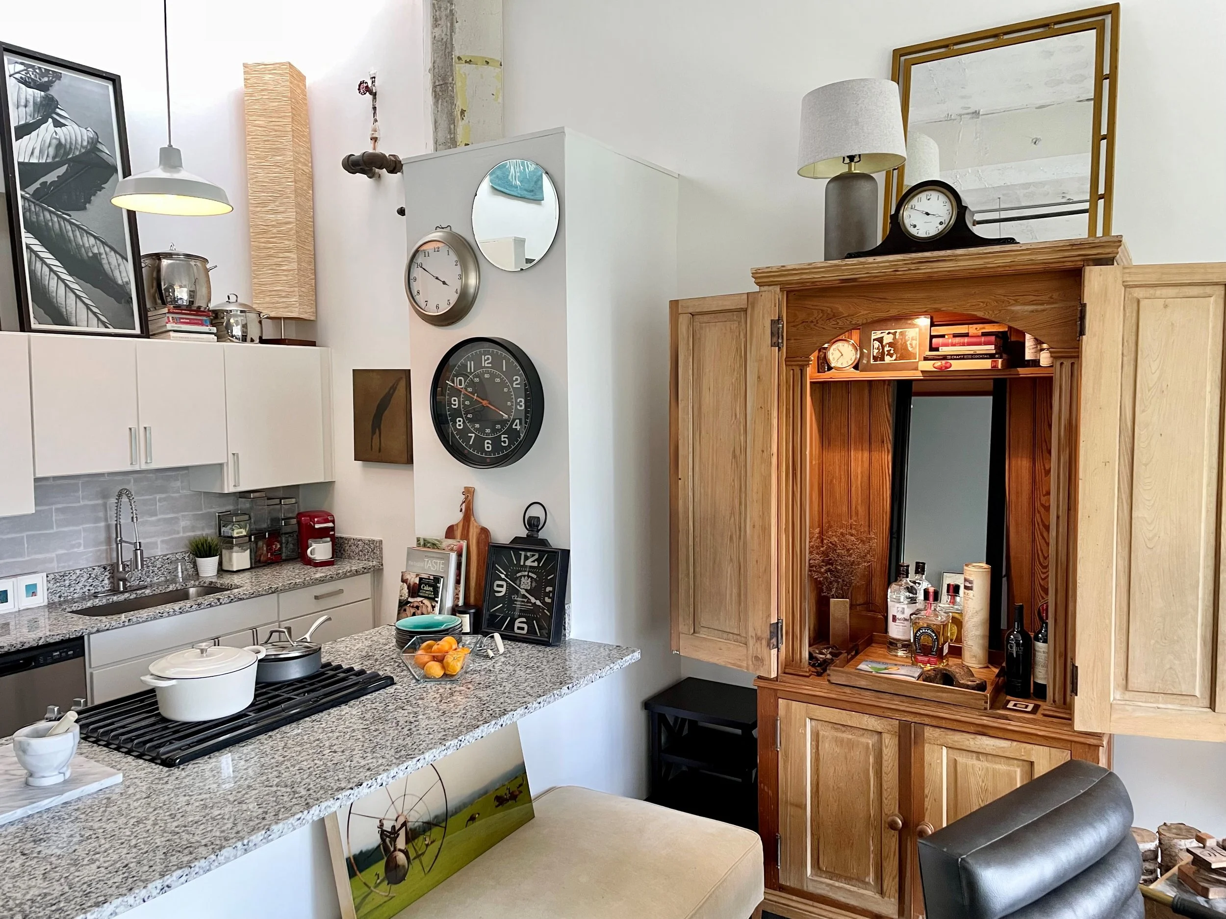 Living room and kitchen area with clocks on the wall, kitchen cabinets, countertop with kitchen items, and a wooden cabinet with a mirror on top.