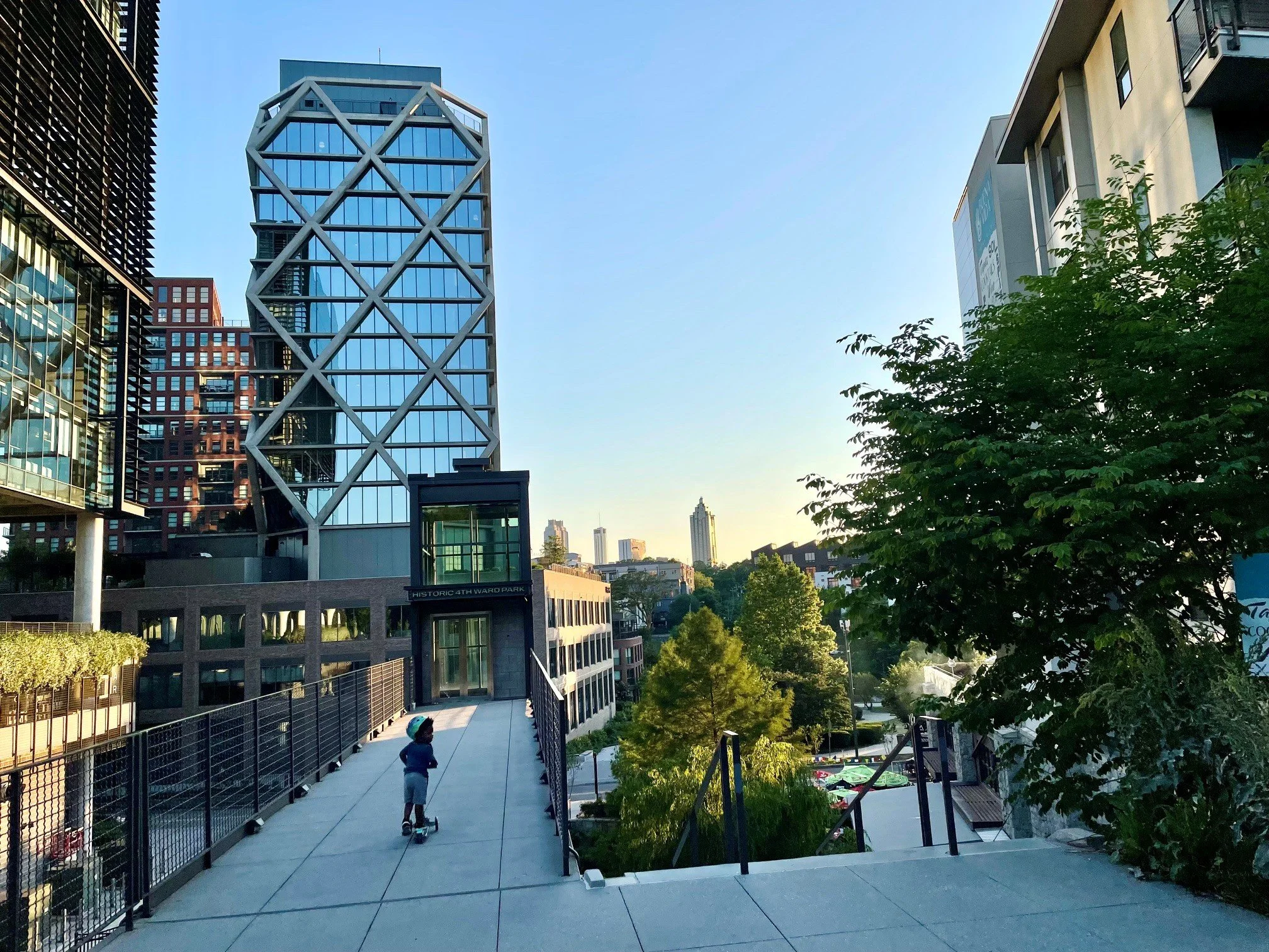 A cityscape with modern high-rise buildings, a pathway with children walking, and trees, during clear weather in an urban park setting.