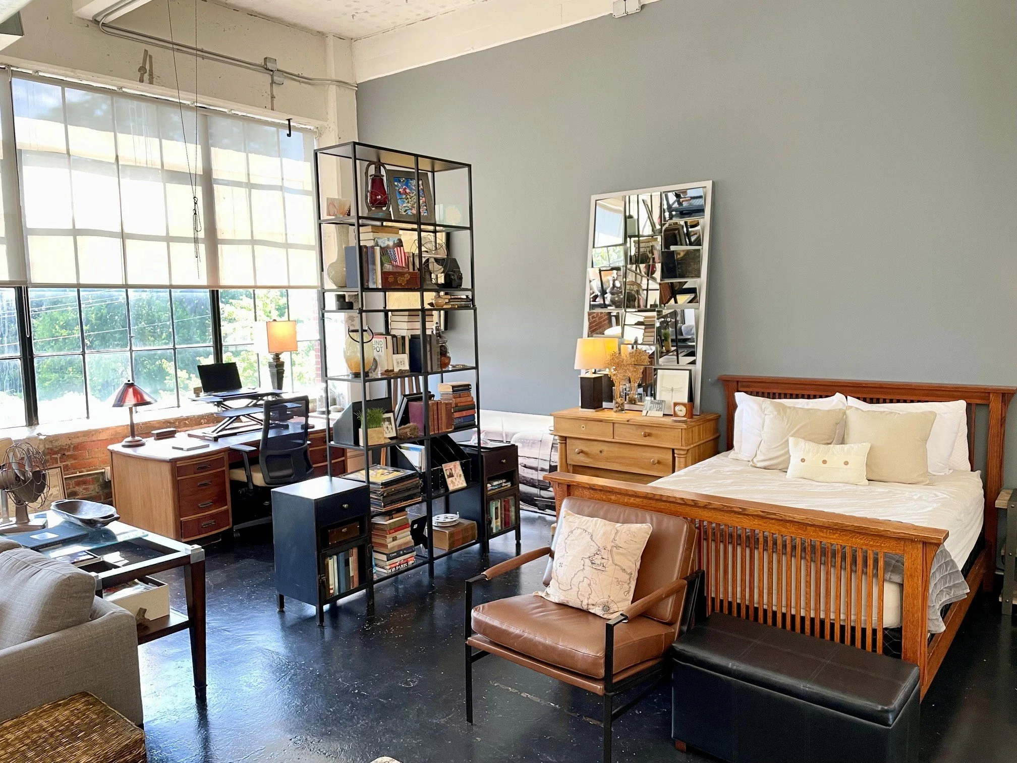 Interior of a bright, modern loft with large windows, a bed with white bedding, a wooden dresser, a leather armchair, a black metal bookshelf filled with books and decorative items, a desk with a lamp, and multiple mirrors reflecting the room.
