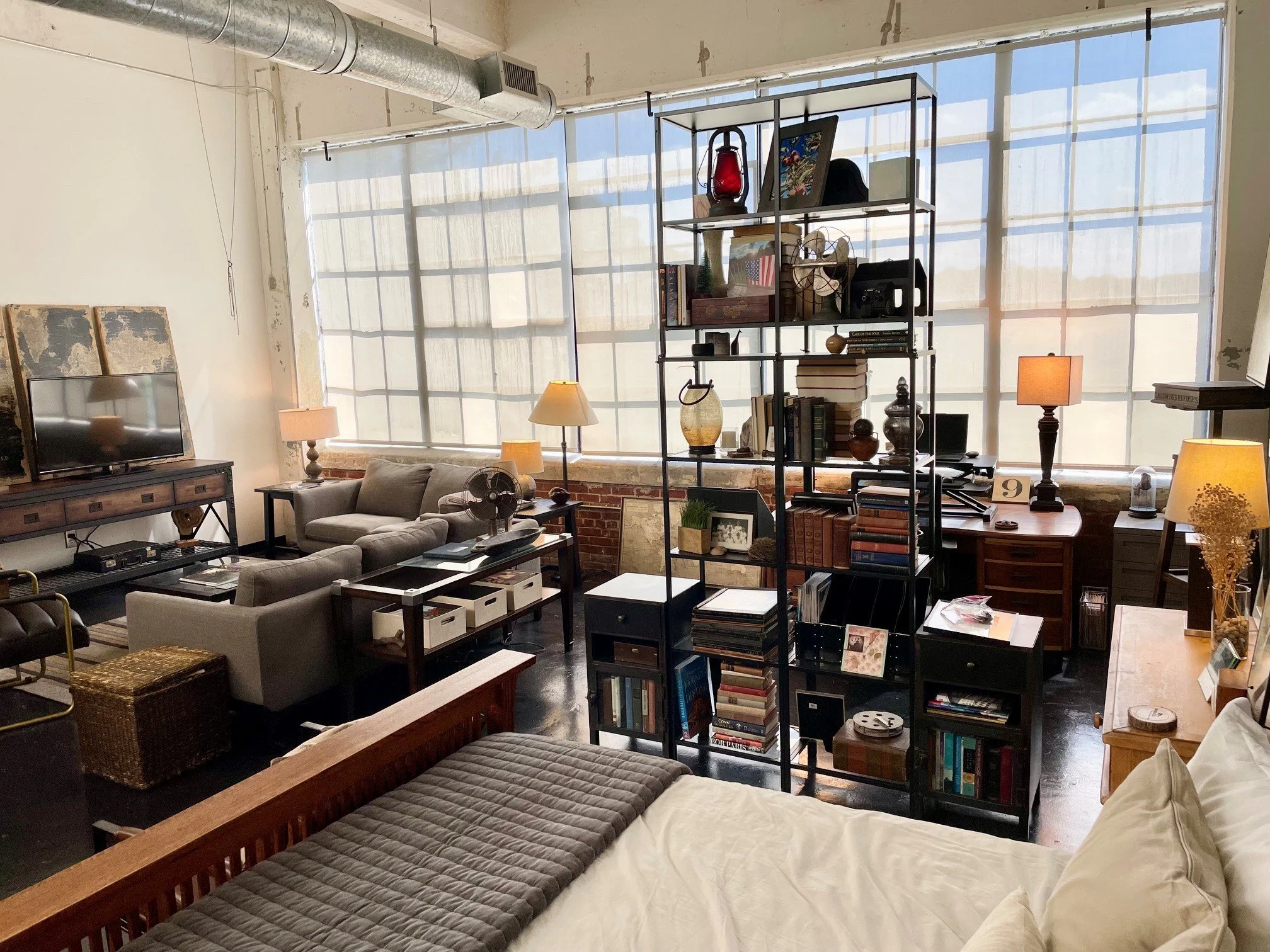 Interior of an apartment with large windows, furniture, shelving unit with decor, and various items.