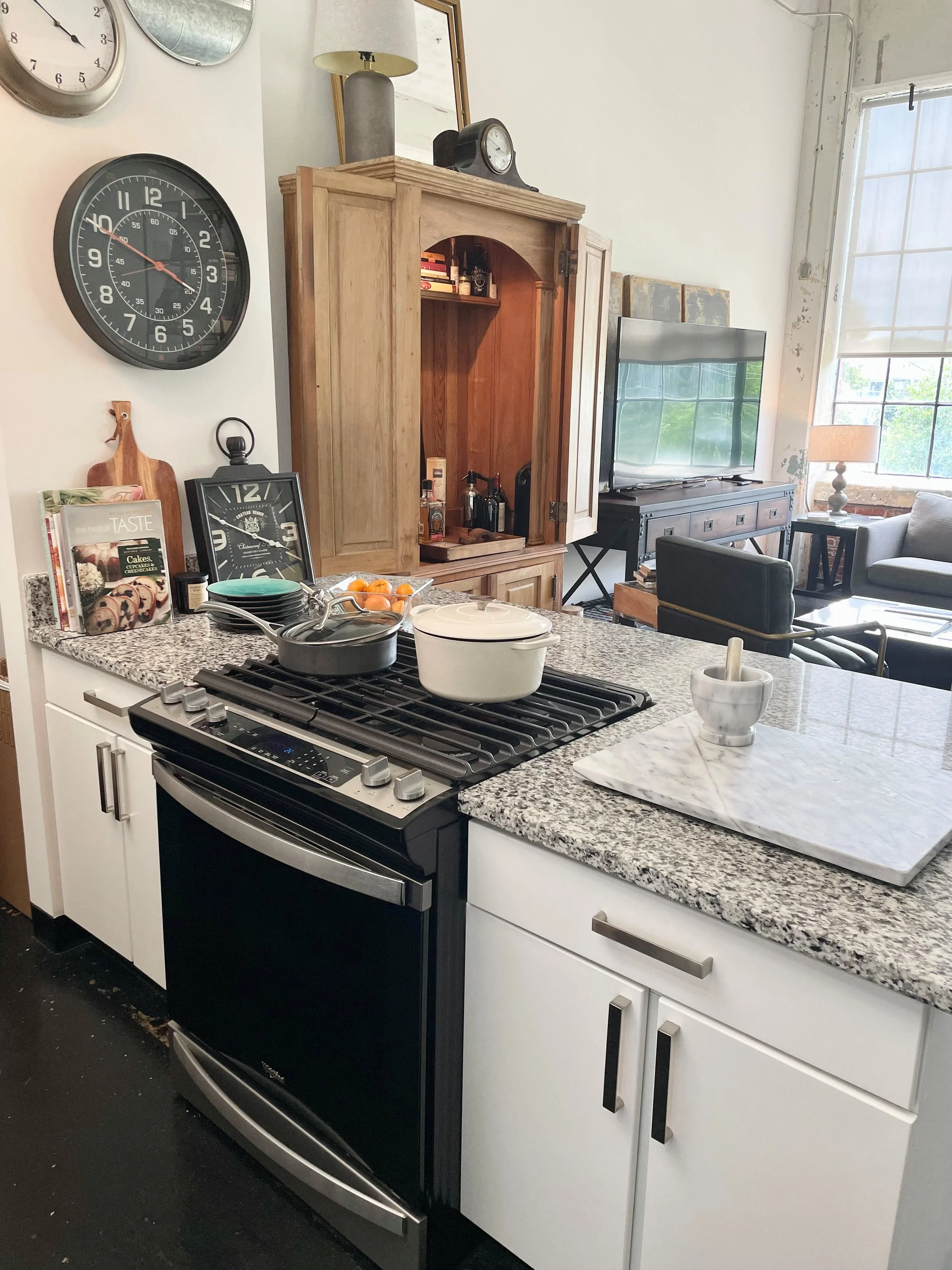 Kitchen with a gas stove, white cabinets, granite countertop, and various kitchen items. In the background, a living room is visible with a TV, a wooden cabinet, and a large window letting in natural light.