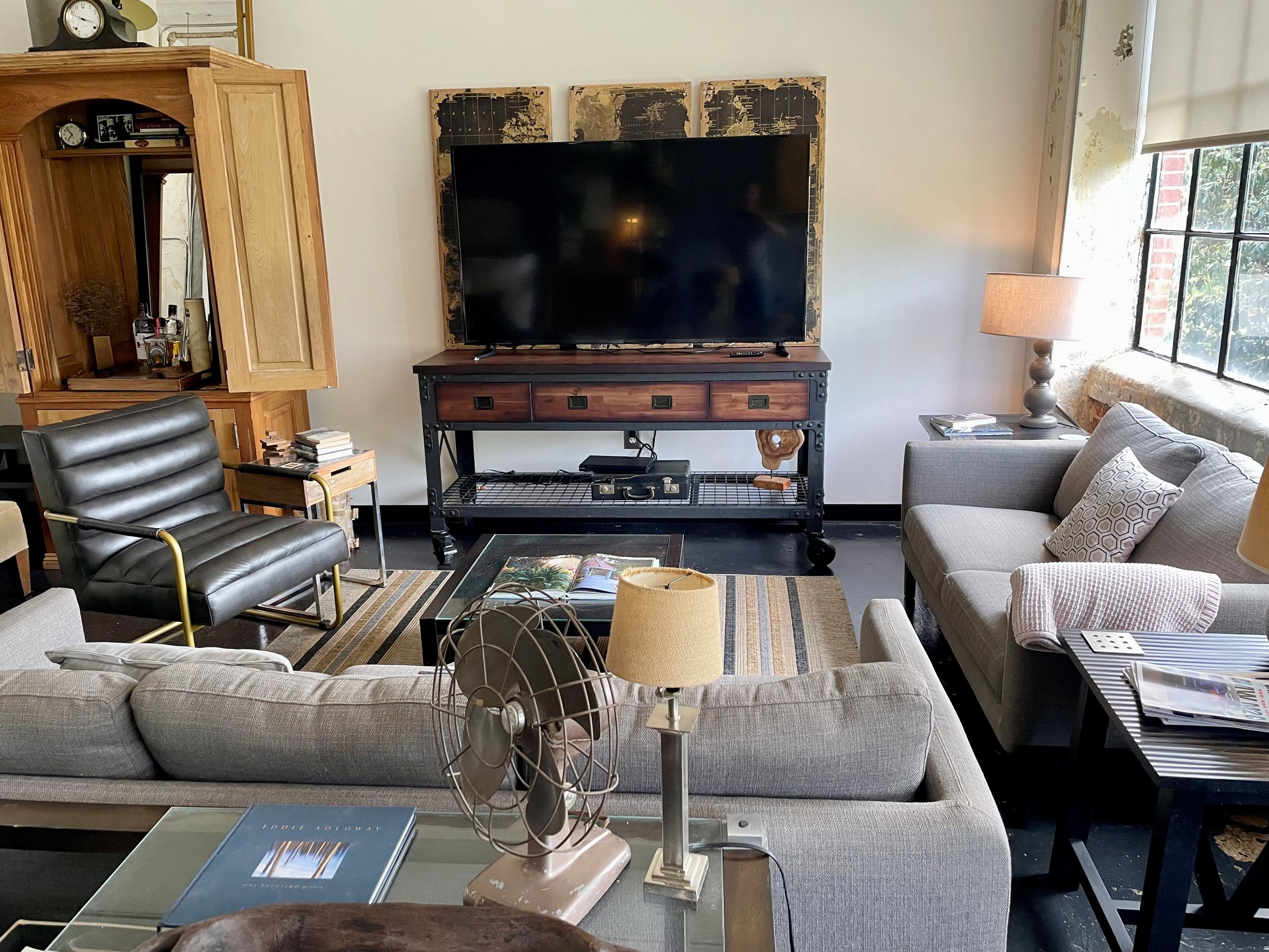 Living room with a gray sofa, black leather armchair, wooden cabinet, large flat-screen TV, side table with lamp, window with partially rolled-up blinds, and decorative elements including books, a vintage fan, and artwork.