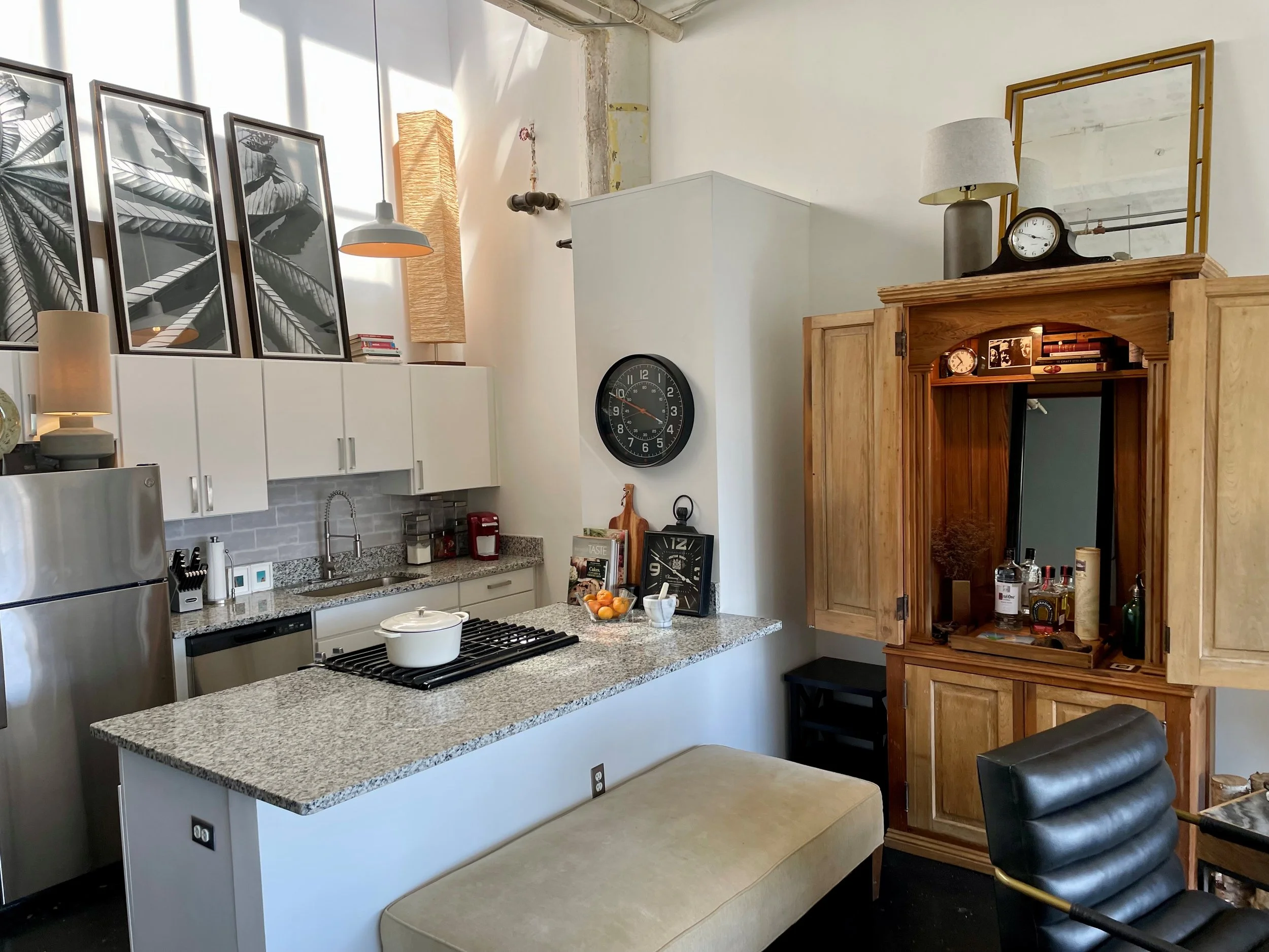 Interior view of a modern kitchen with white cabinets, granite countertops, and several framed black-and-white botanical prints on the wall. There is a pot on the stove, a bowl of oranges, and various kitchen appliances. To the right, a wooden armoir