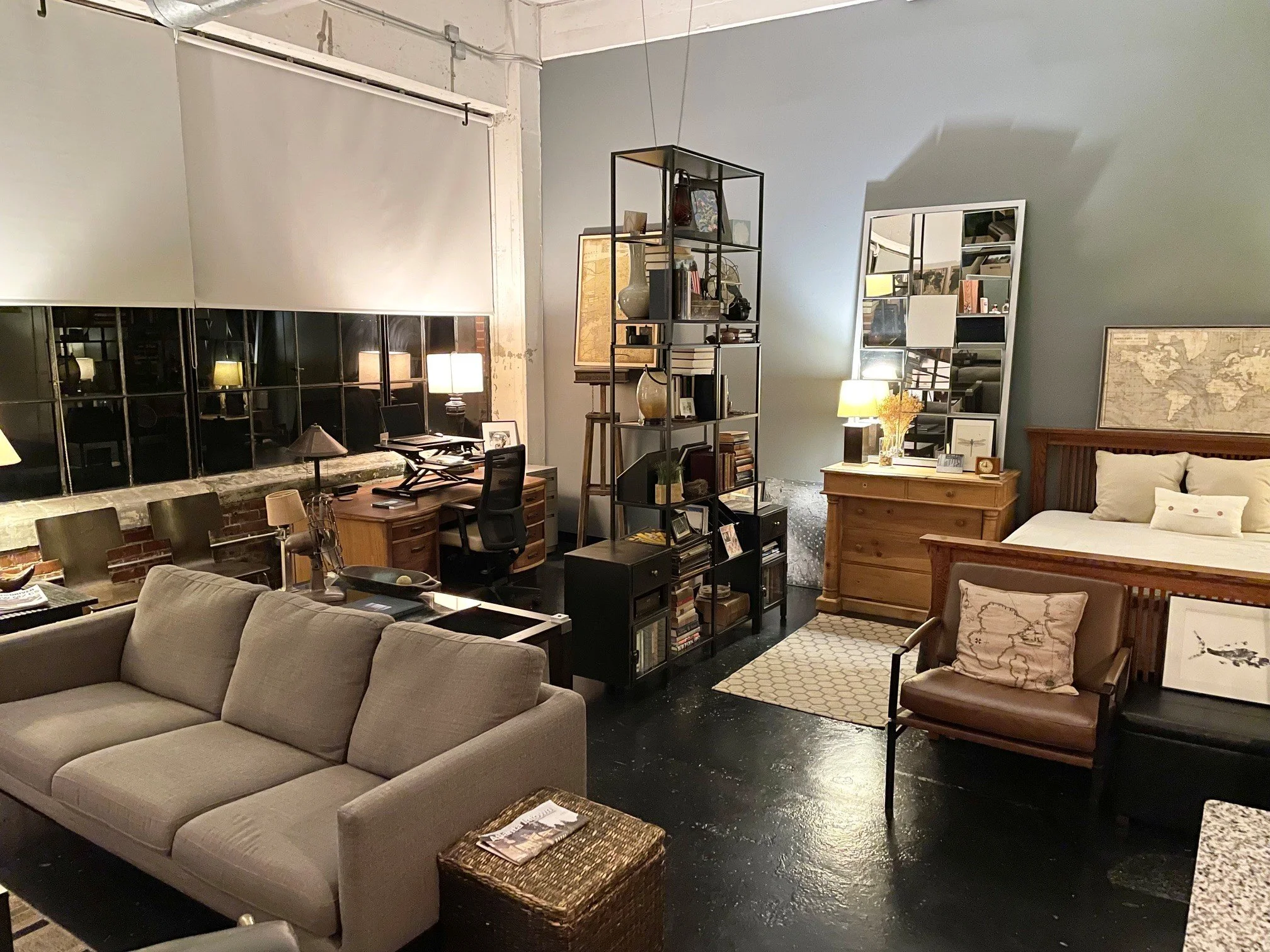 Several seating areas including a beige couch and a leather armchair with a pillow, a wooden dresser with a lamp and framed pictures, a black shelving unit with books and decorative items, a large mirror, and multiple lamps providing warm lighting in