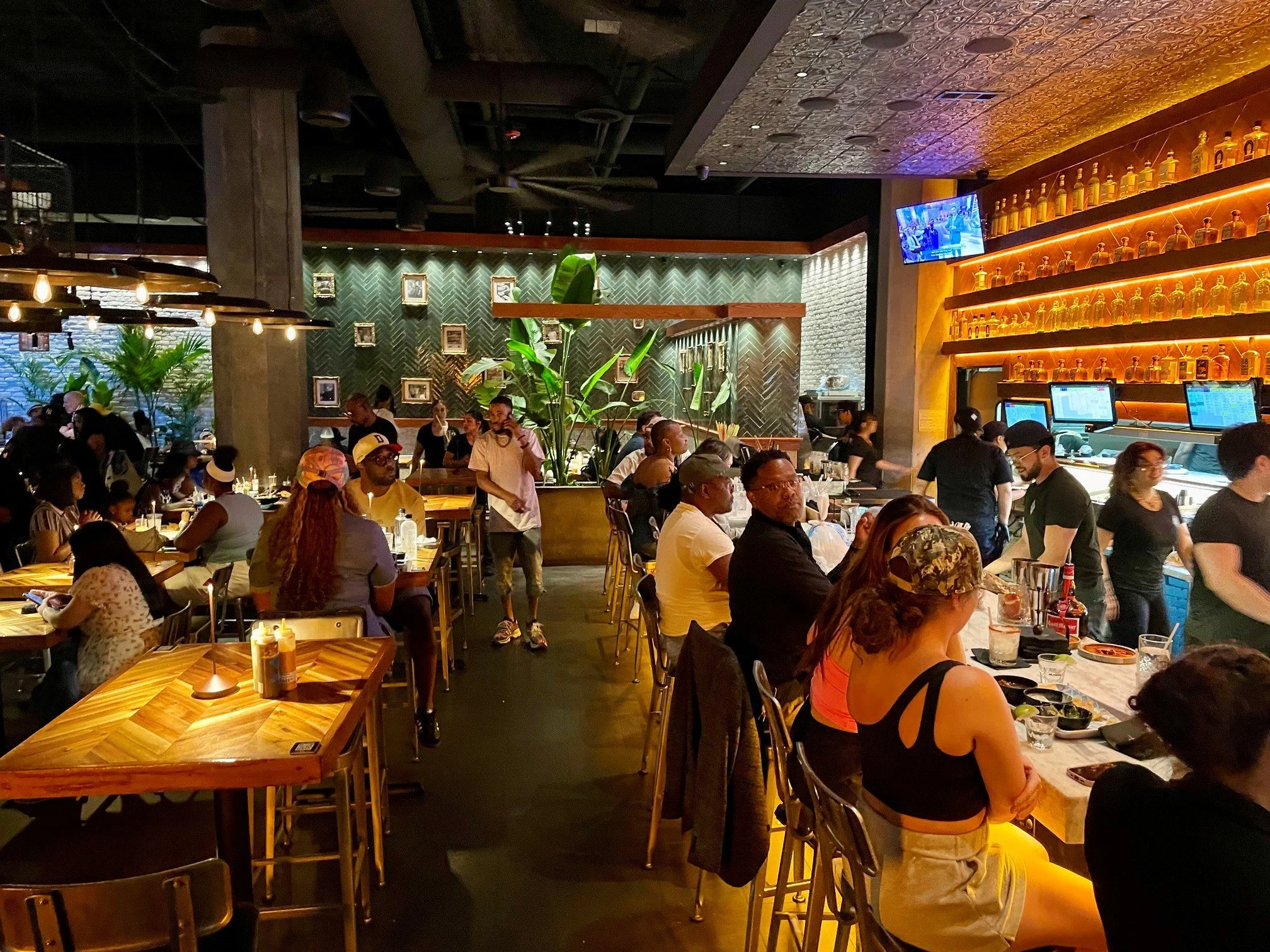 A busy restaurant with many people dining. There are tables with food and drinks, plants for decoration, a patterned green wall with picture frames, and a bar area with bottles and a TV.