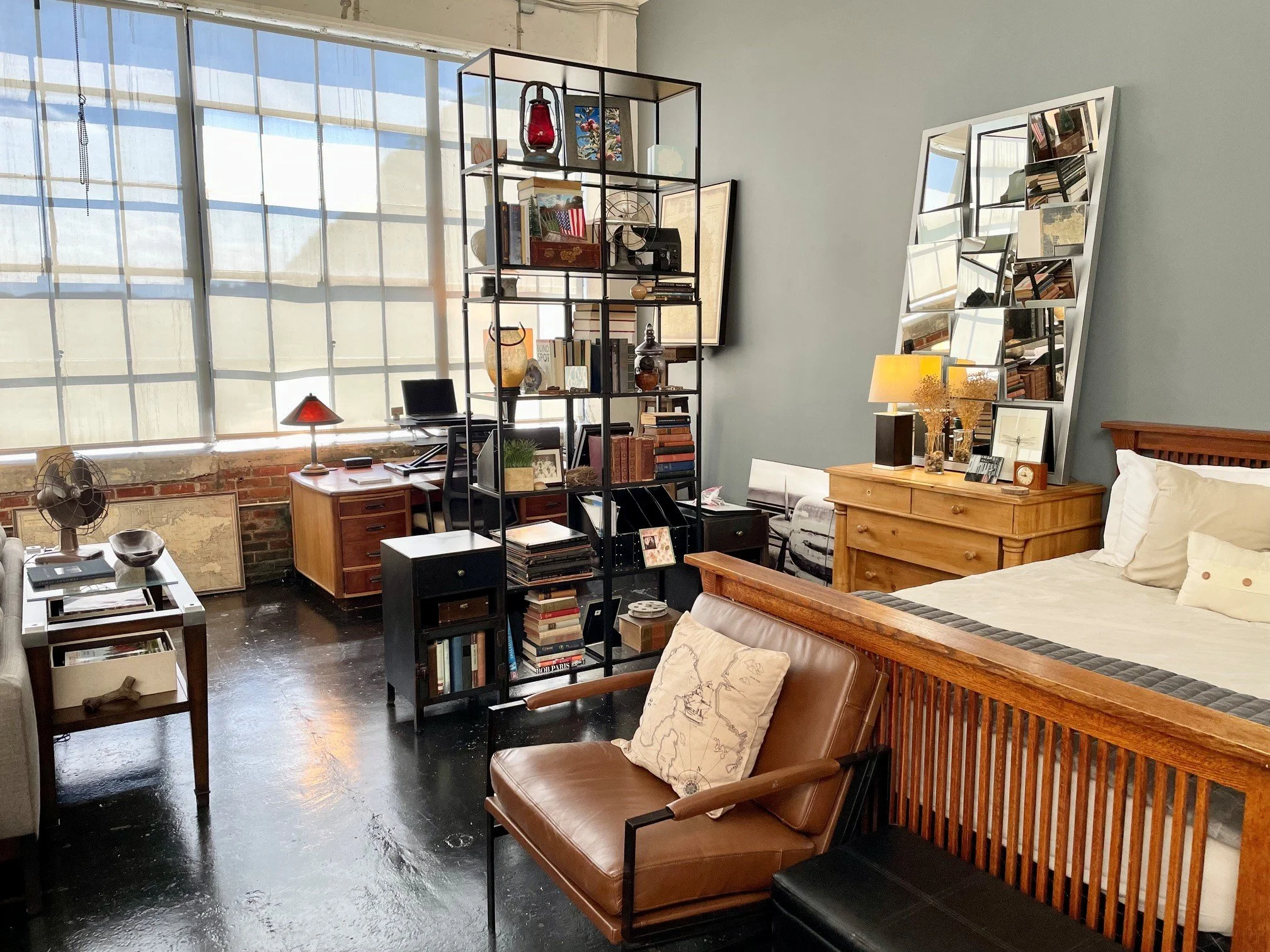 A cozy living space with large windows, a black metal bookshelf filled with books and decorative items, a wooden dresser with a lamp, a small desk with a computer, a leather armchair with a map pillow, a large mirror with a geometric frame, and a bed with white bedding. The room has a mix of modern and rustic decor with exposed brick and wood accents.