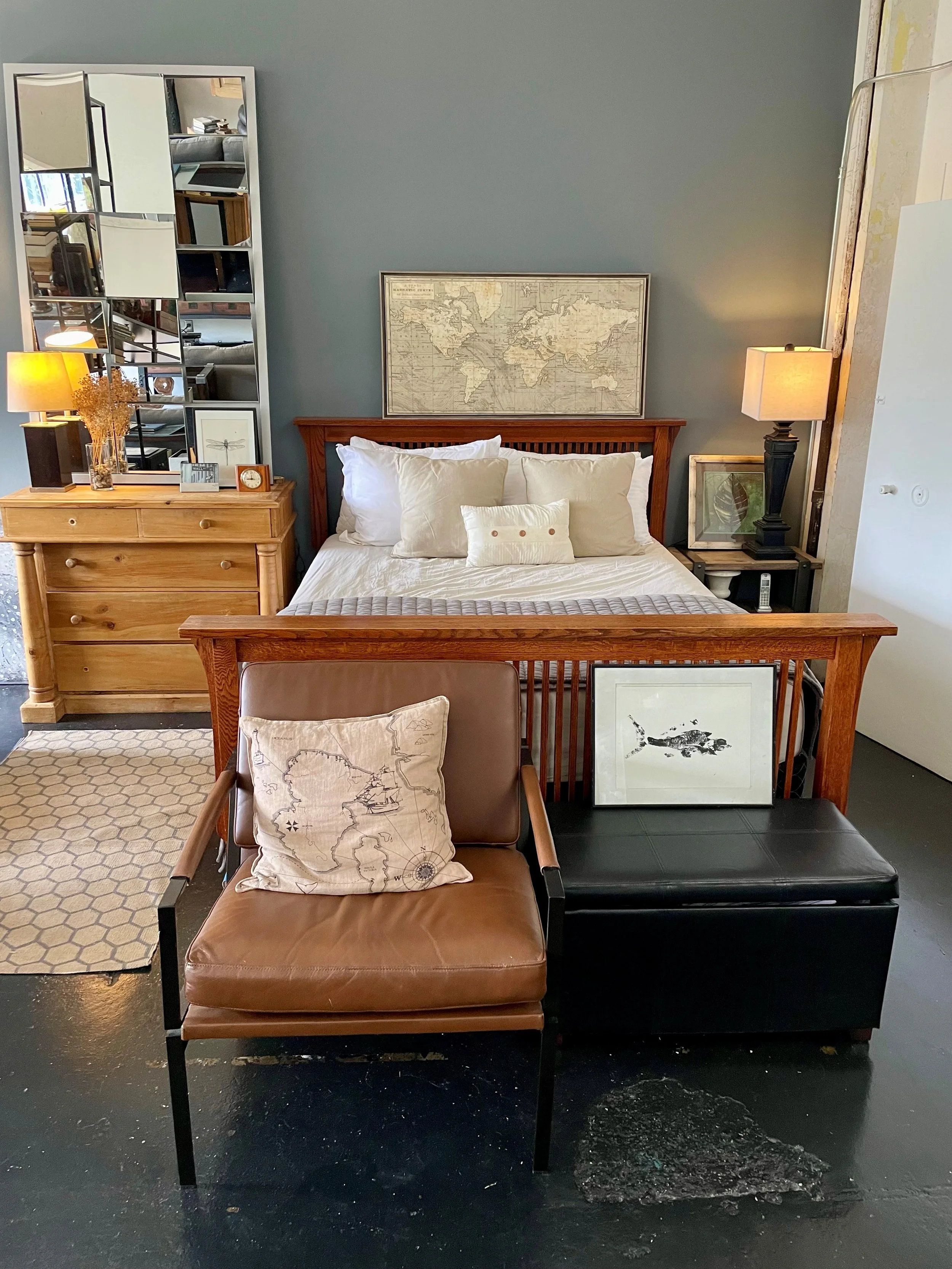 Living room with a wooden bed frame, white bedding, and multiple pillows, flanked by two nightstands with lamps. A large mirror with a geometric design hangs on the left wall, and artwork including a world map and a fish print are visible, along with