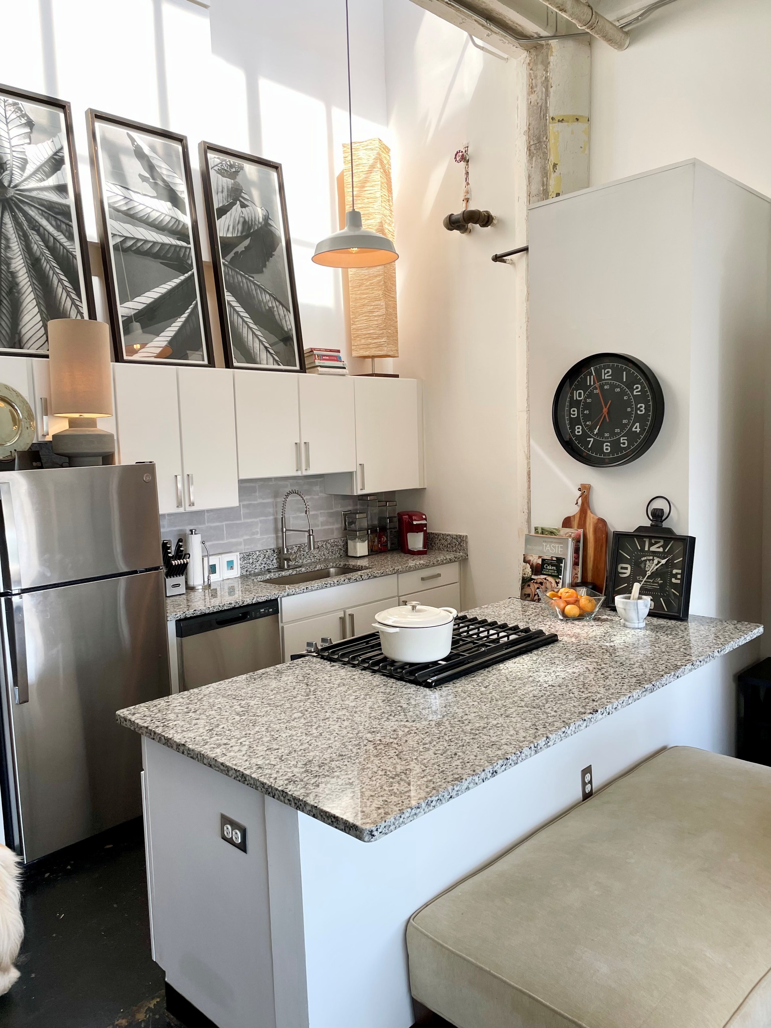 A modern kitchen with white cabinets, a granite countertop, a stove with a white pot, and decorative wall art. There are clocks, magazines, and a bowl of peaches on the counter.