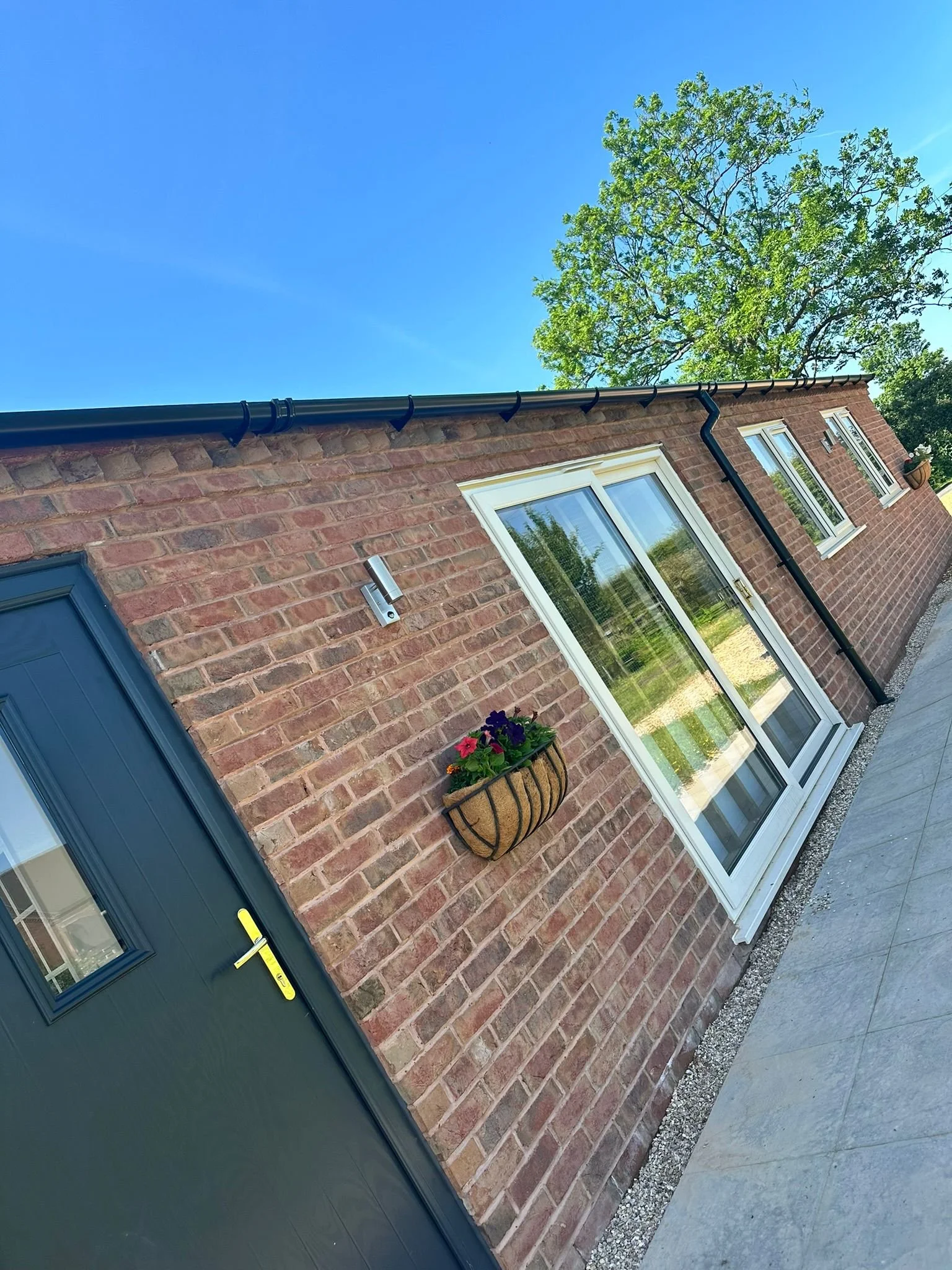 Brick house exterior with a blue door, windows, a hanging flower basket, and a black drainpipe under a clear blue sky, with a tree in the background.
