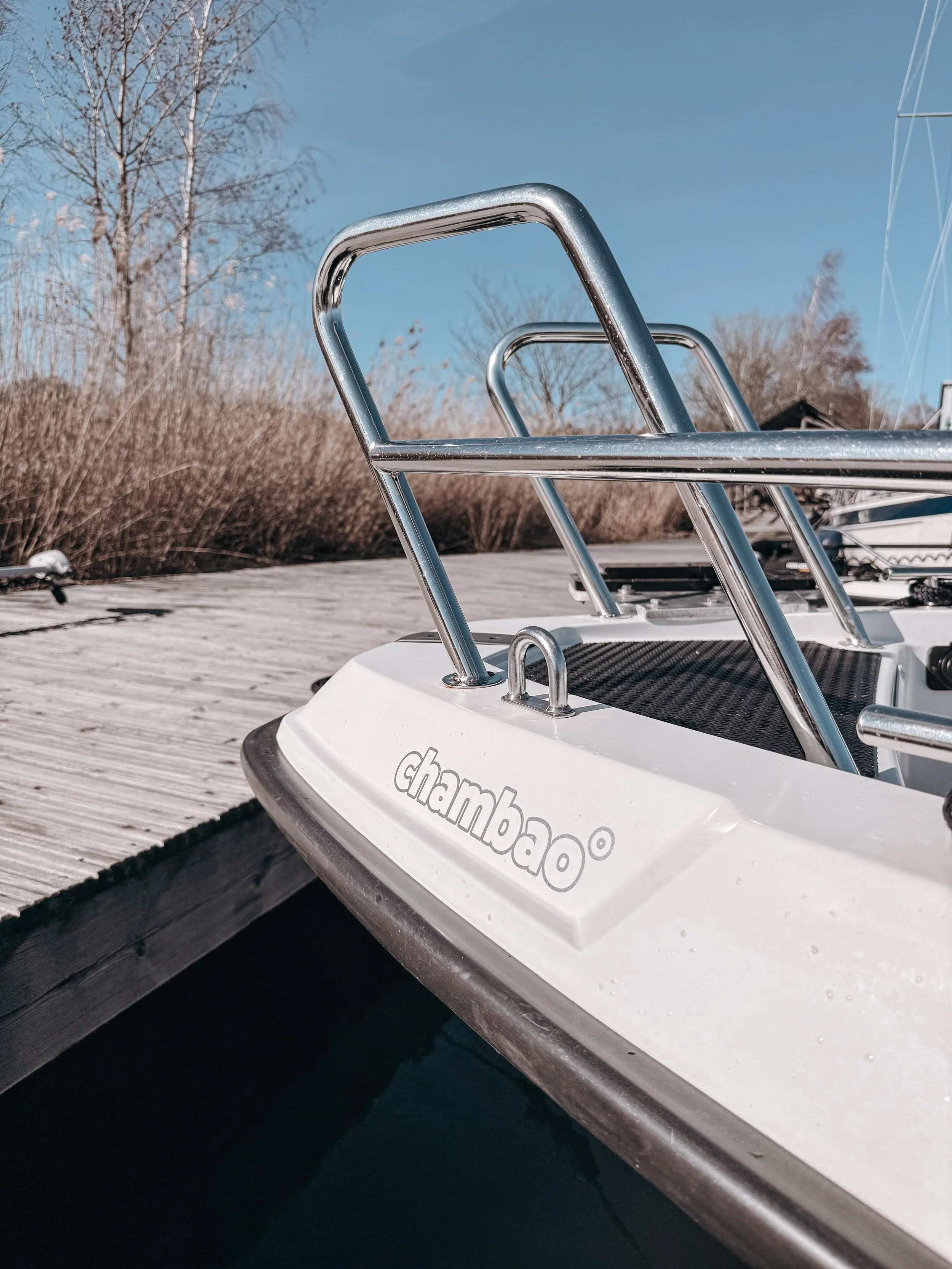 Close-up of a white boat with the word 'chambao' written on the side, docked at a wooden pier, with a clear blue sky and leafless trees in the background.