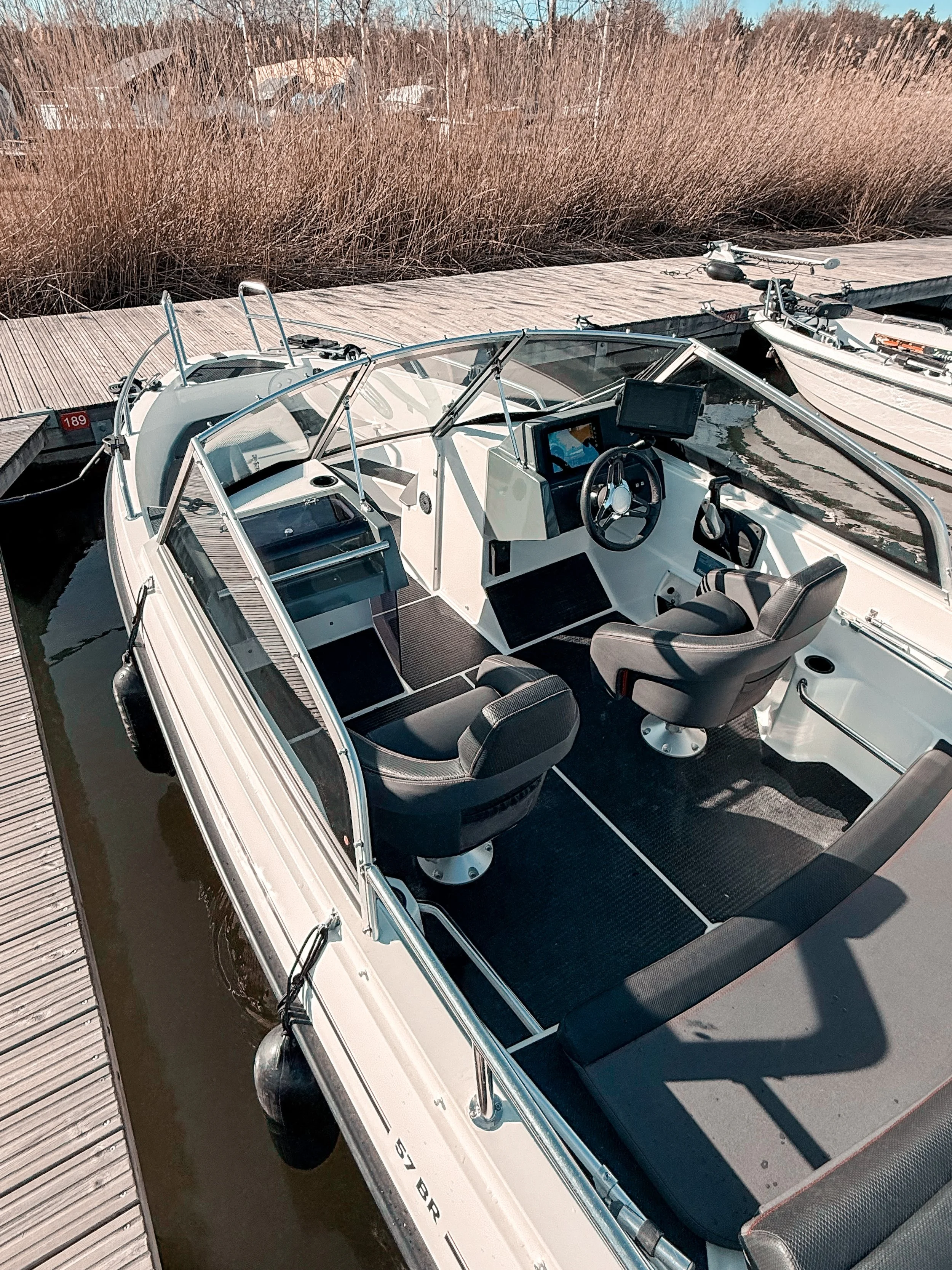 A white boat docked at a marina with a brown wooden pier, with tall brown grasses and houses in the background, and another similar boat nearby.