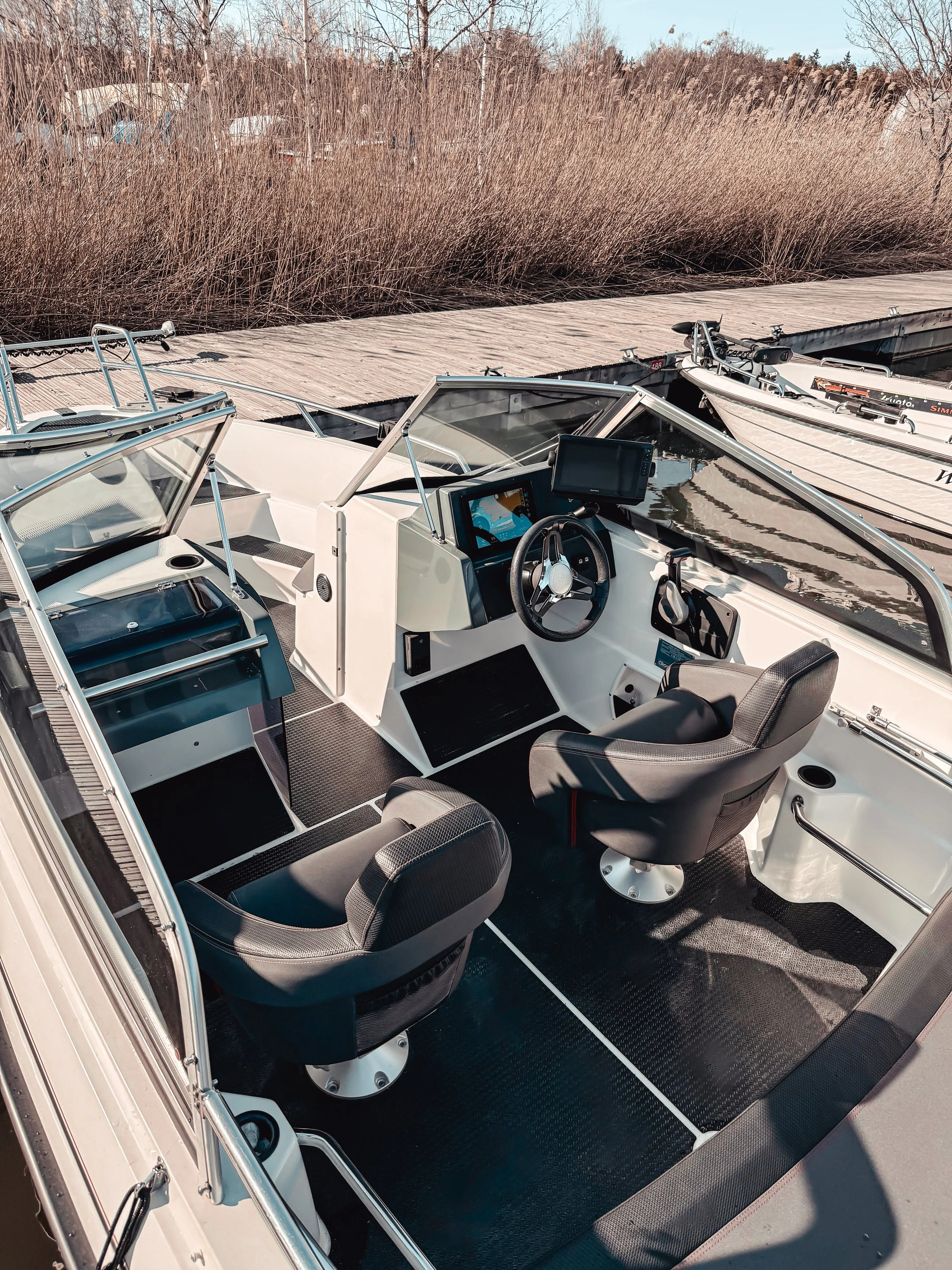 View of a boat interior with steering wheel, dashboard, and two black seats, docked at a boat dock with tall reeds and houses in the background.