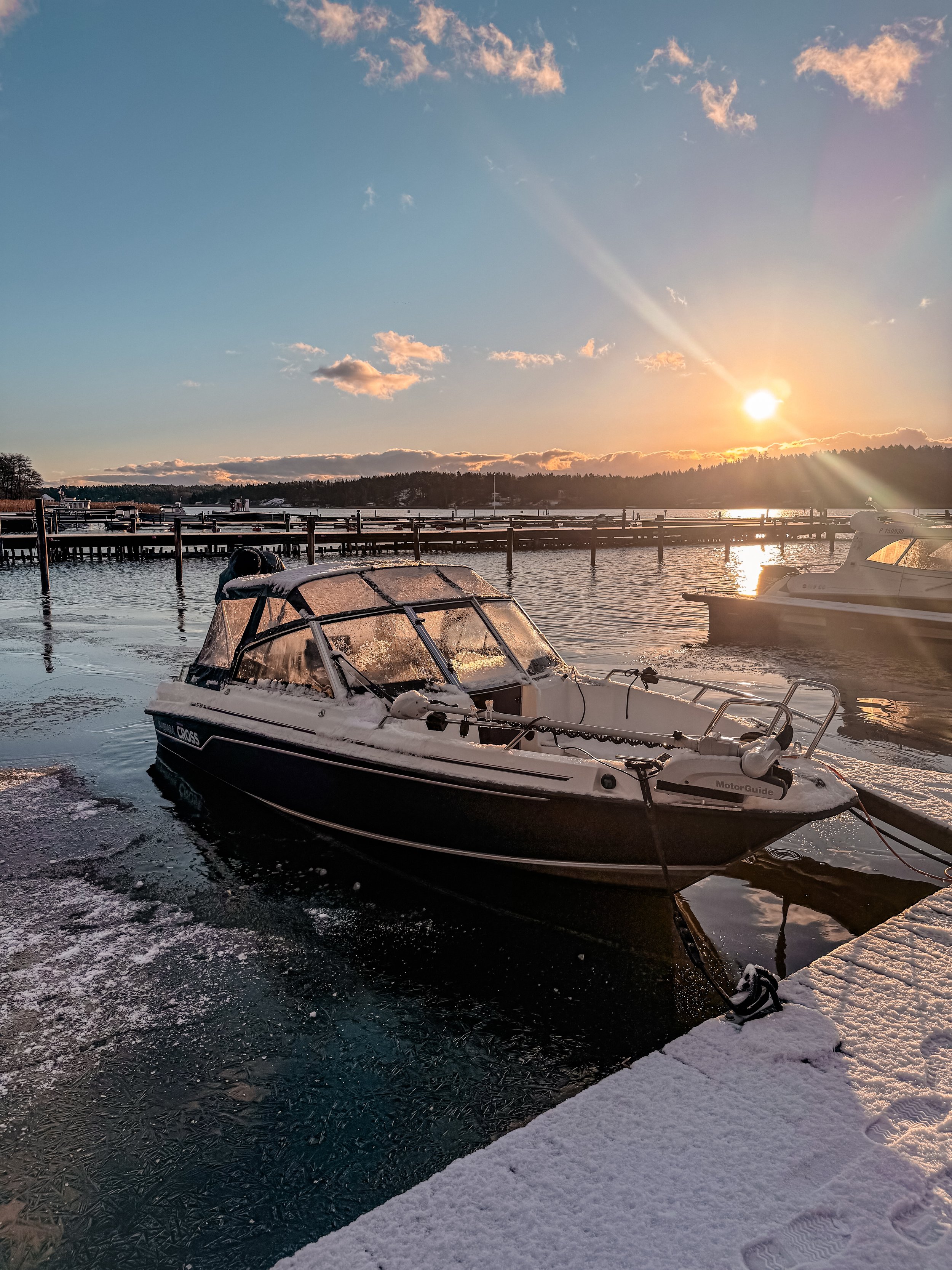 A boat docked at a snowy shoreline during sunset, with a calm body of water and a forest in the background.