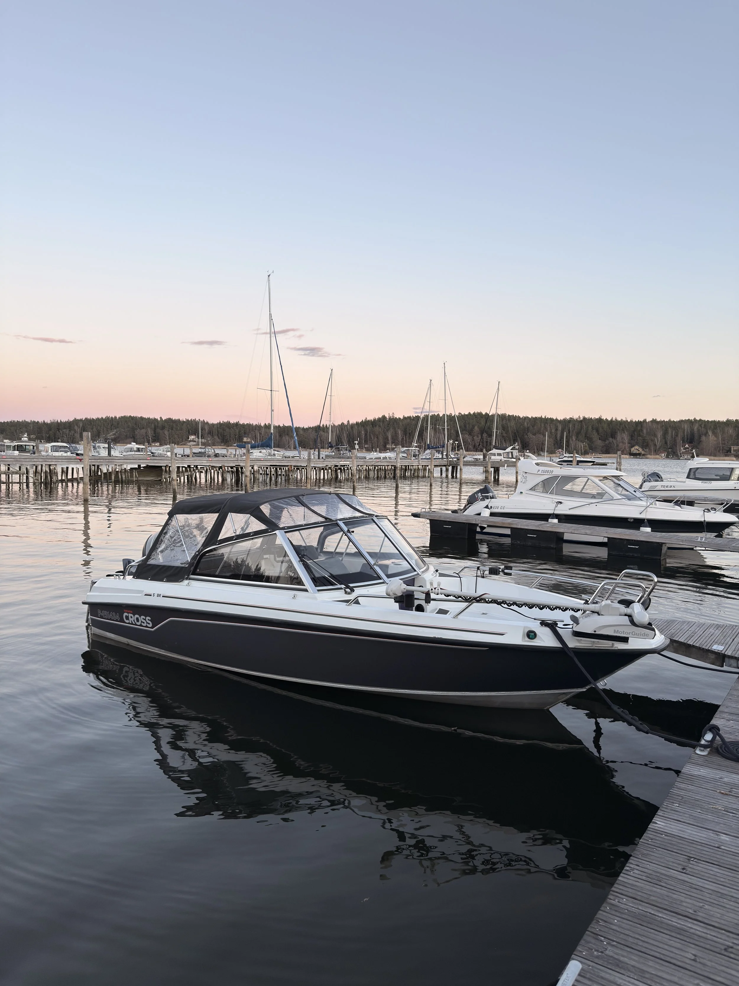 Boats docked at a marina during sunset with calm water reflecting the boats and a wooden dock on the right side.