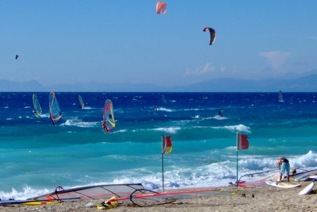 People windsurfing and kiteboarding on the ocean, with a sandy beach in the foreground and a few flags planted in the sand.