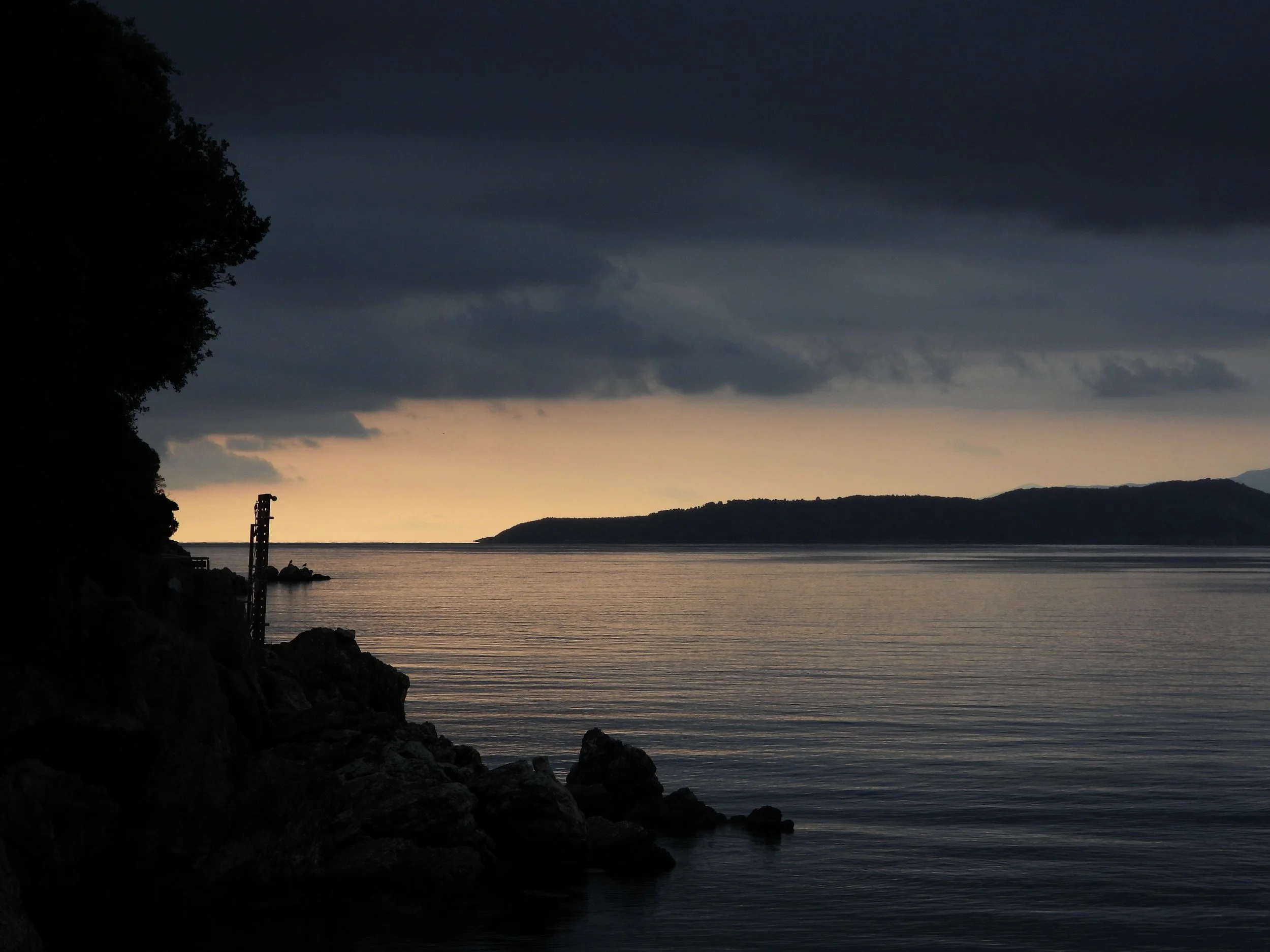 A lakeside scene at dusk with dark clouds in the sky, a calm body of water, a silhouetted forested shoreline, and a small weather station or similar structure in the foreground.