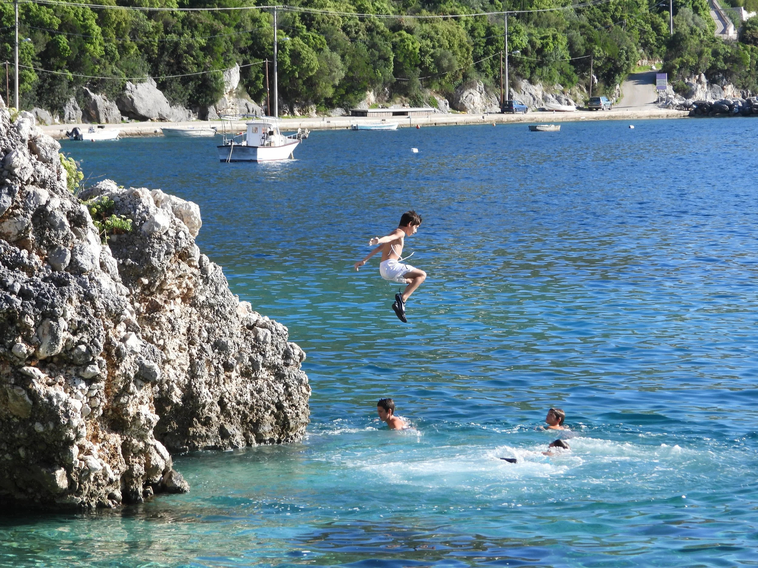 Young boy jumping into the water from rocks on a sunny day, with other children swimming nearby, boats anchored in the water, a tree-covered hillside, and a road with parked cars in the background.