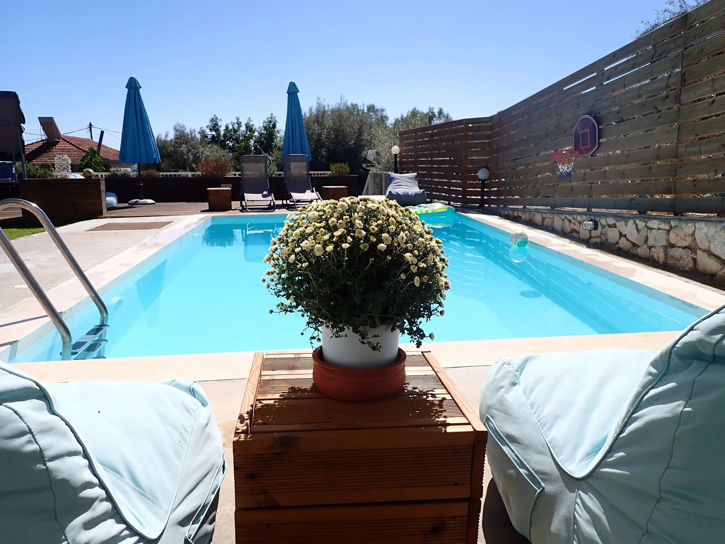 A backyard with a swimming pool, surrounded by lounge chairs, umbrellas, and outdoor furniture, with a wooden deck and a privacy fence.