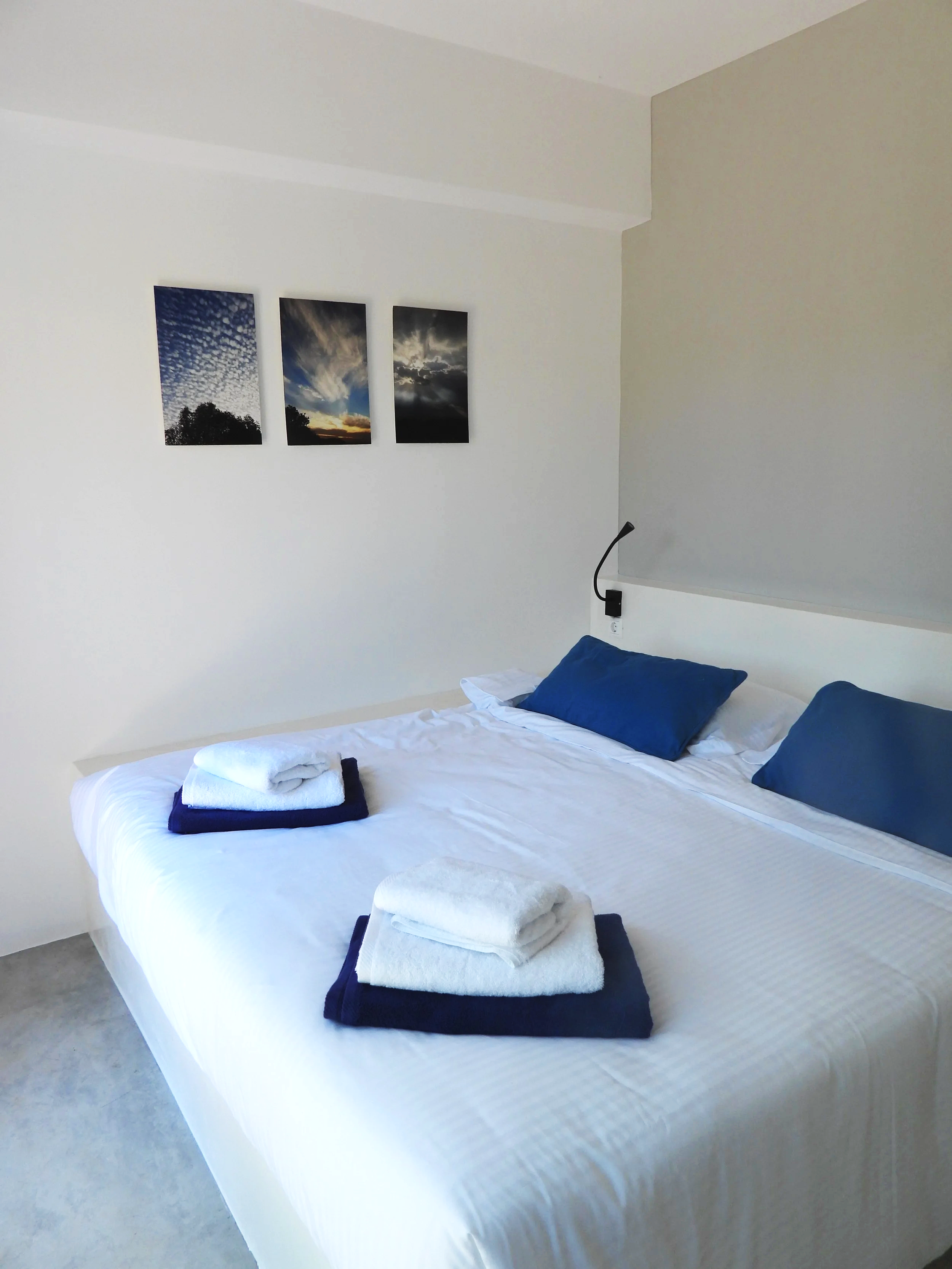 A neatly made bed with white linens, two blue pillows, and folded white towels on dark blue towels at the foot of the bed. Three framed sky and cloud photographs hang on the wall above the bed.