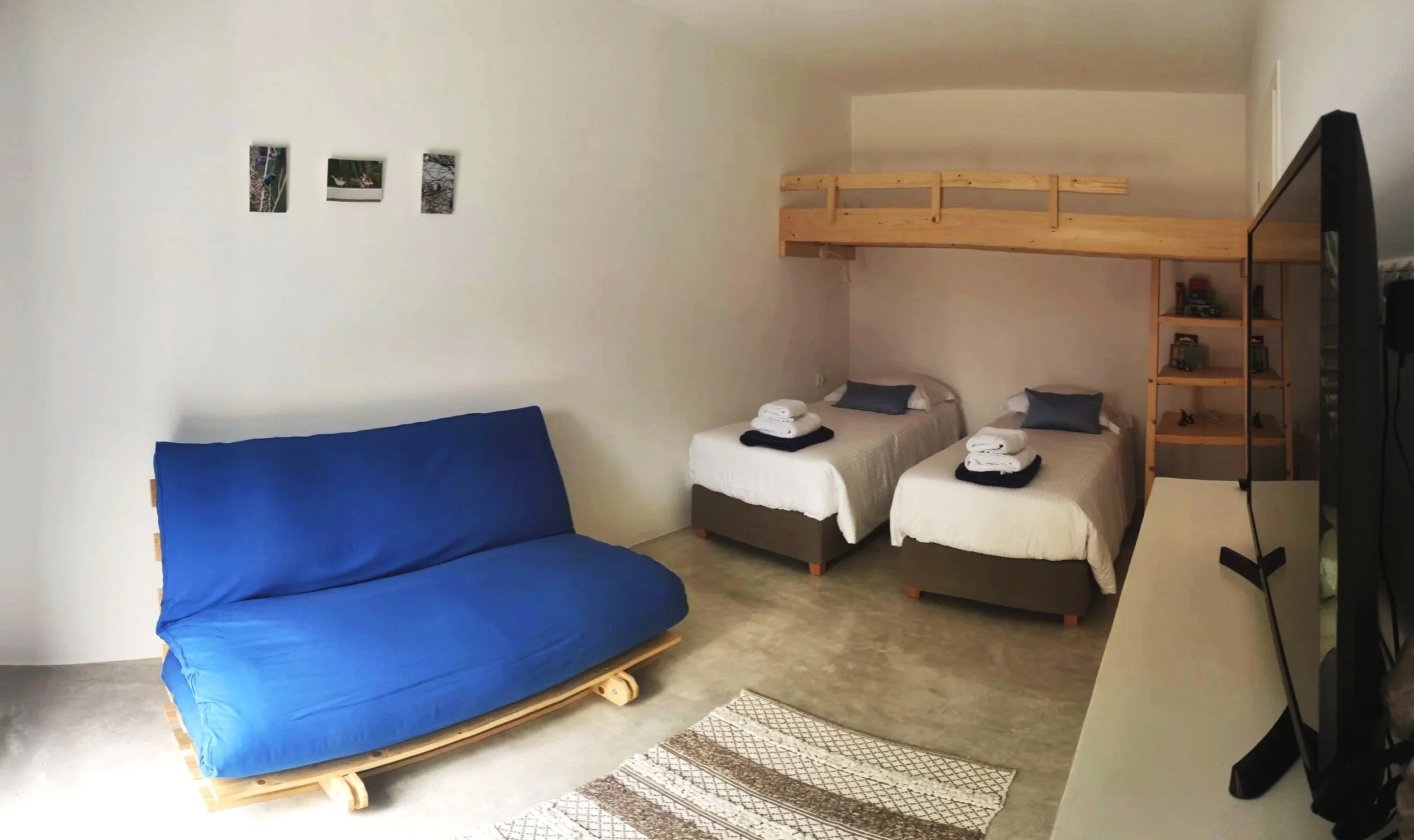 A room with two single beds, each with white bedding and gray pillows, with folded towels on top, a blue couch on a wooden pallet frame, a wooden loft above the beds, a wall-mounted TV, and small framed artwork on the wall.