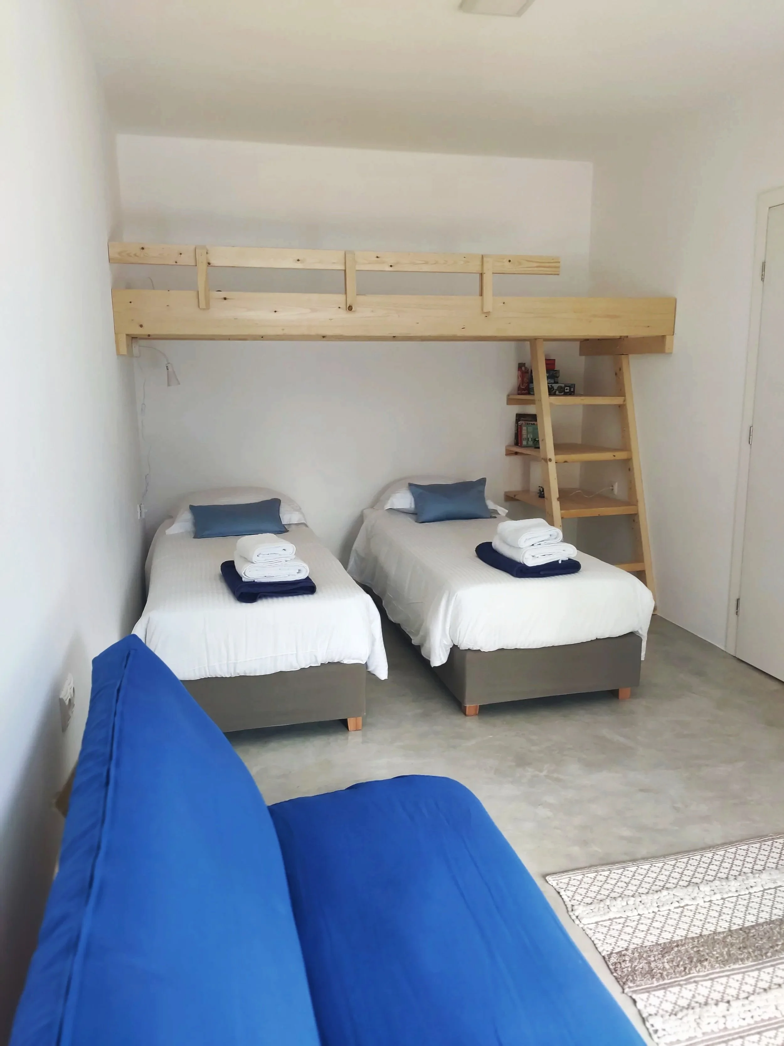The image shows the perspective from a person lying on a blue couch, looking towards a room with two single beds each with white sheets, a blue pillow, and a stack of towels at the foot. There is a wooden loft bed over the beds with a ladder and smal