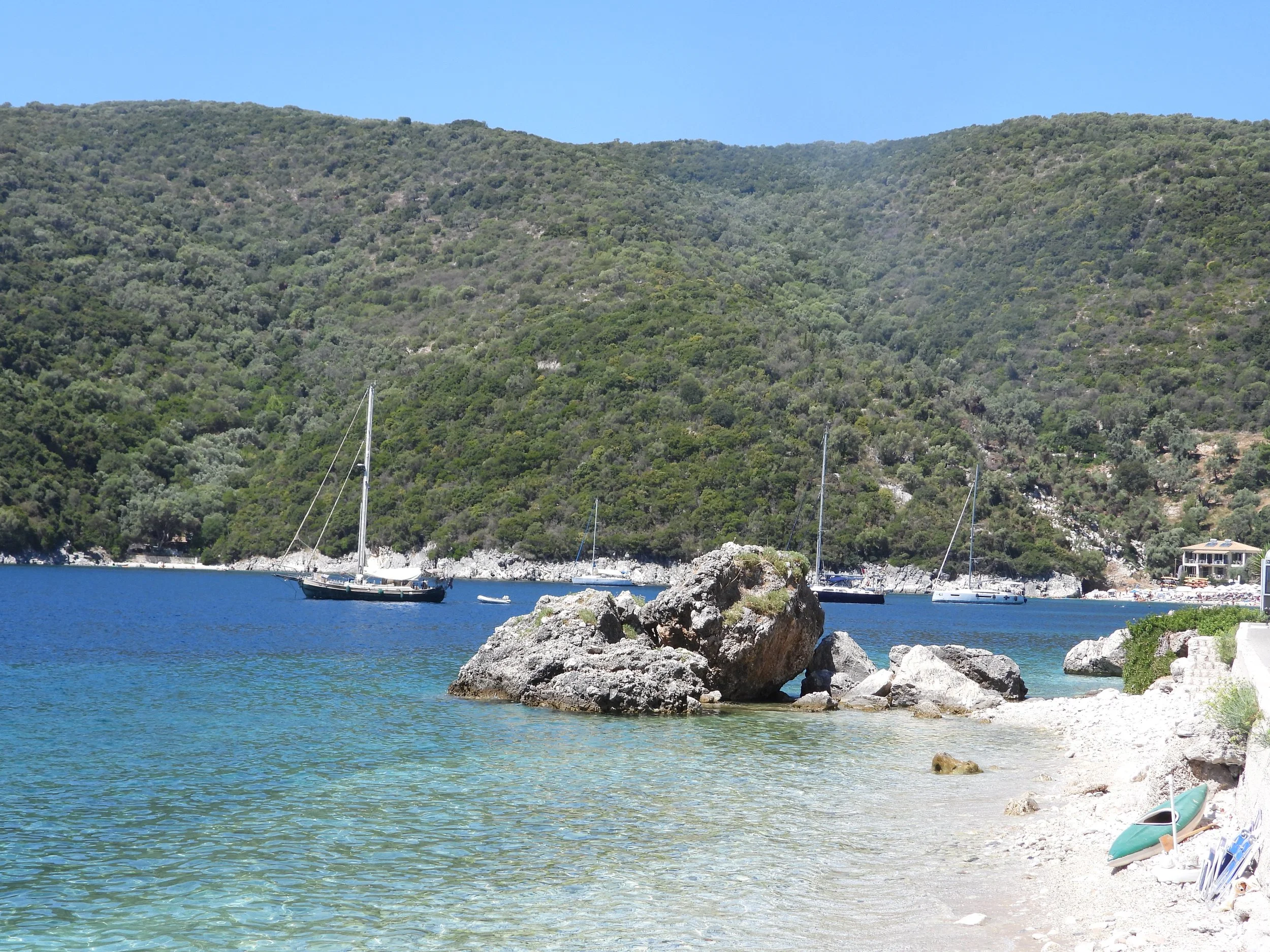 A coastal scene with clear blue water, rocks in the foreground, sailboats with masts, a hillside covered with trees, and some buildings along the shoreline under a blue sky.