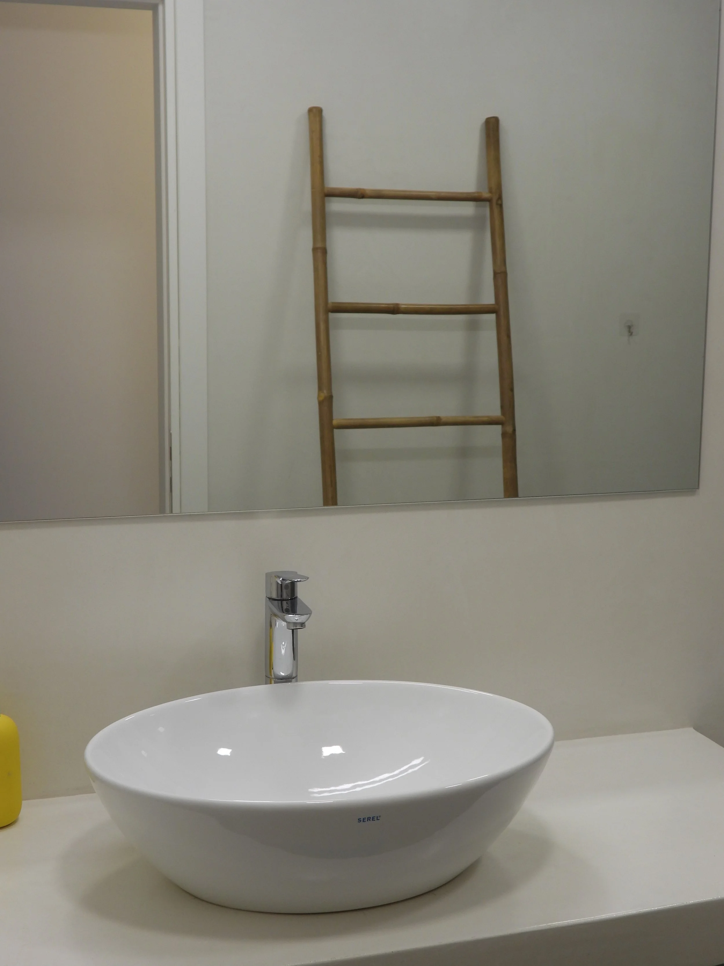 A bathroom sink with a white oval basin, a silver faucet, a mirror on the wall reflecting a wooden ladder, and a yellow object on the left side of the counter.
