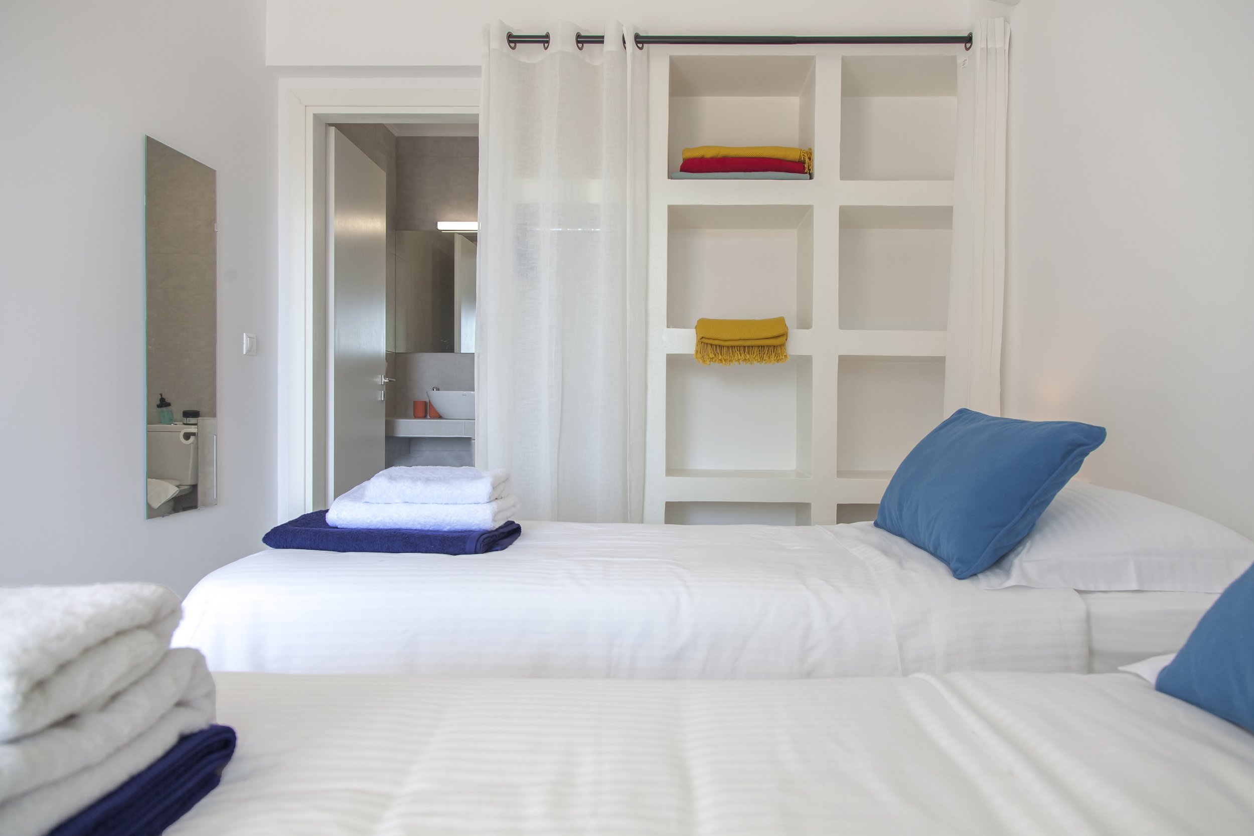 A bedroom with a bed featuring a blue pillow, white sheets, and folded towels on the bed. There is a large mirror on the wall, an open doorway leading to a bathroom with a sink and orange accents, and a white shelf with folded towels and a yellow clo