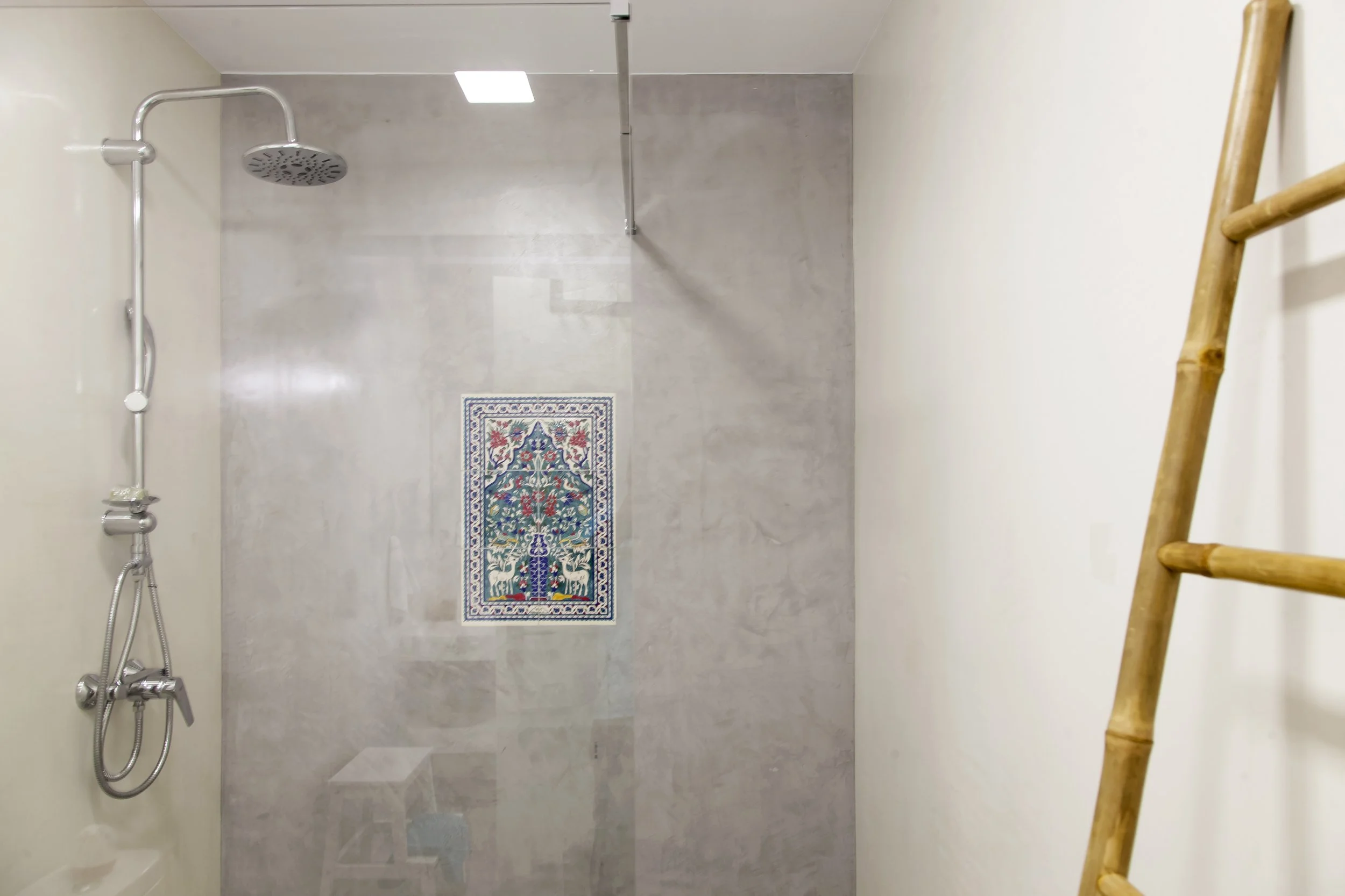 A minimalist walk-in shower with beige tiled walls and floor, a black and white patterned bath mat, a silver rain showerhead, and a bamboo ladder leaning against the side wall.