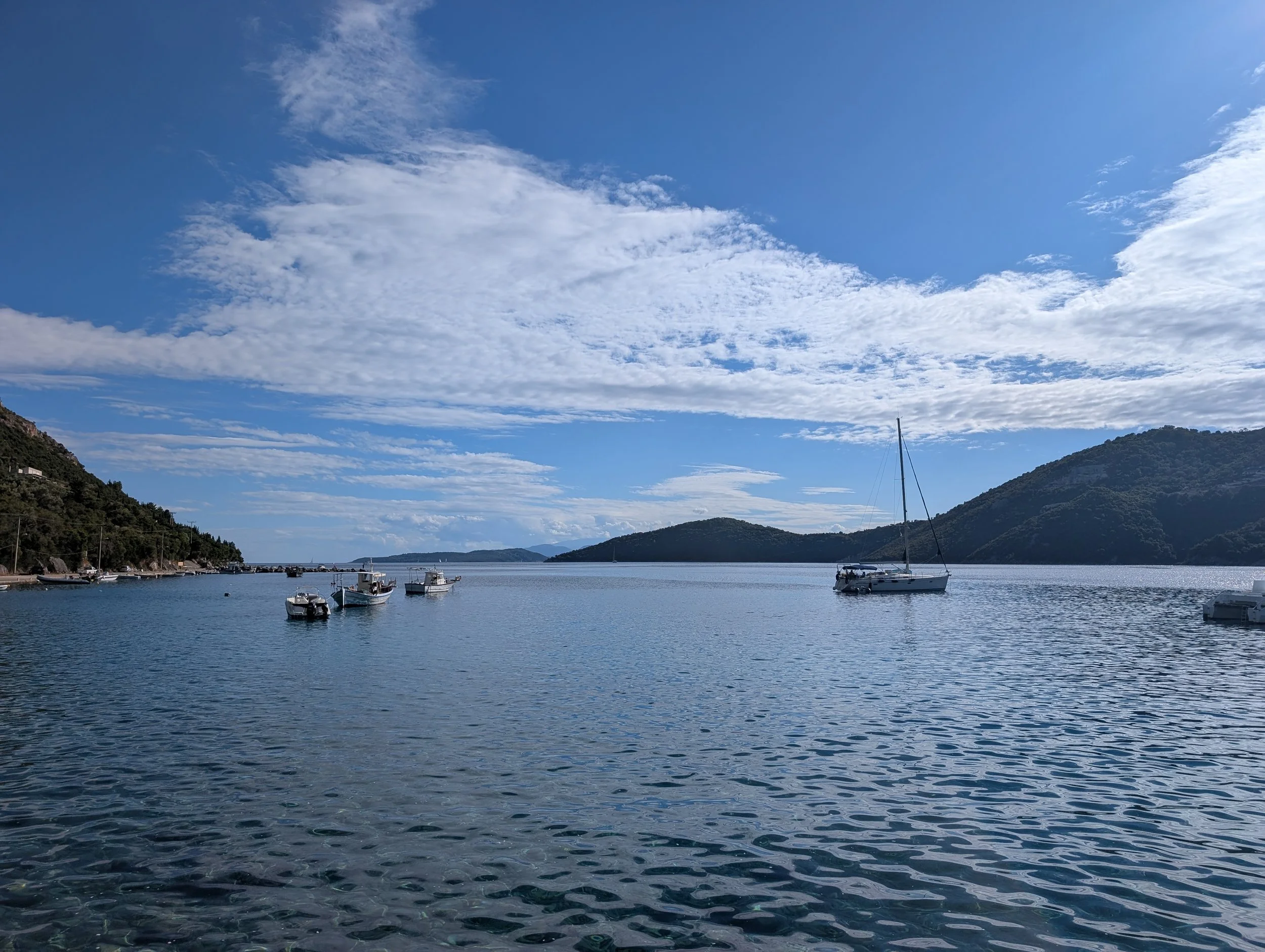 A harbor with multiple boats floating on calm water, surrounded by green hills and mountains under a partly cloudy blue sky.