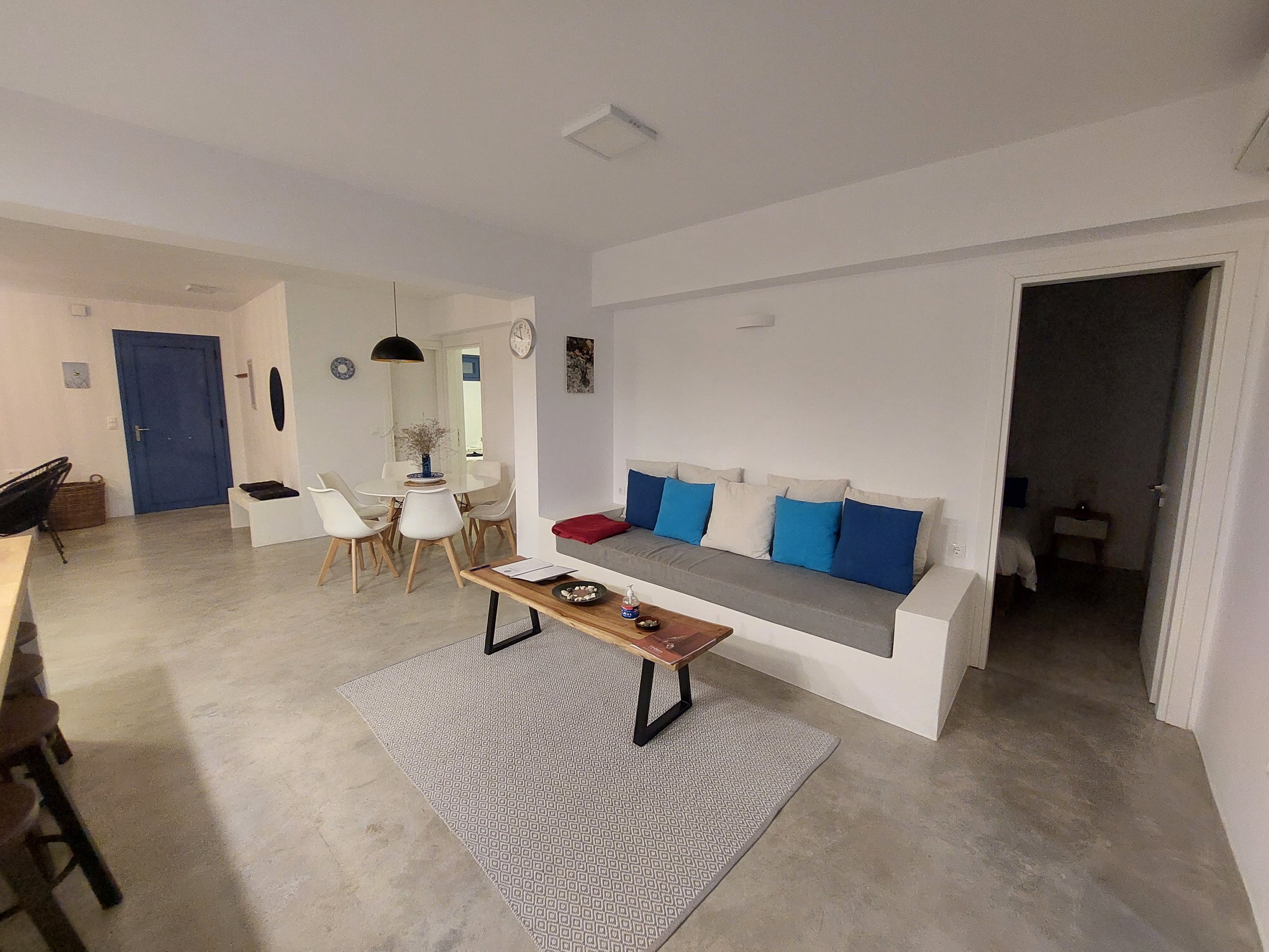 Living room with white walls, a blue front door, white dining table with six chairs, a black pendant light, a wall clock, and a gray sofa with blue and white cushions.