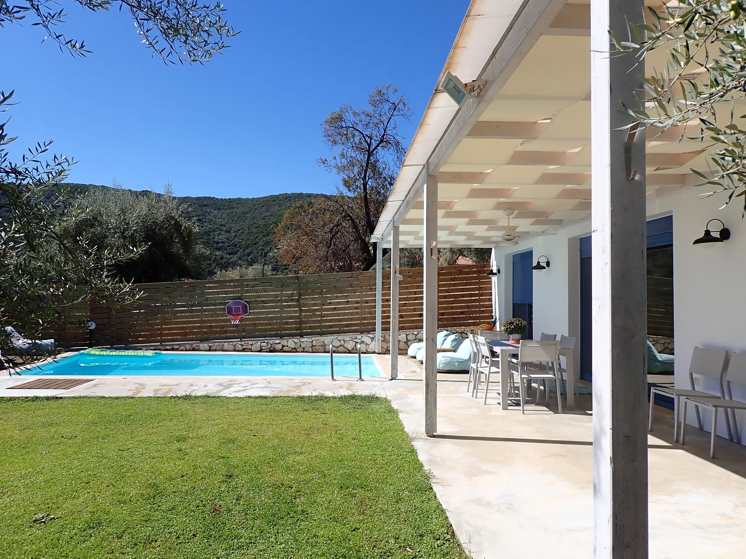 A backyard with a swimming pool, a wooden fence, and a patio area with chairs and tables under a covered porch. There is a basketball hoop near the pool and trees surrounding the area, with hills in the background and a clear blue sky.