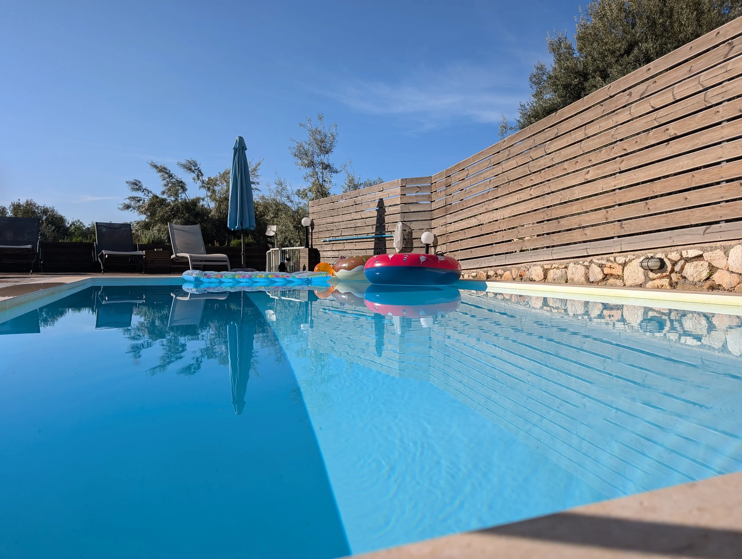 Swimming pool with inflatable toys, lounge chairs, a blue umbrella, and a wooden privacy wall under a clear blue sky.