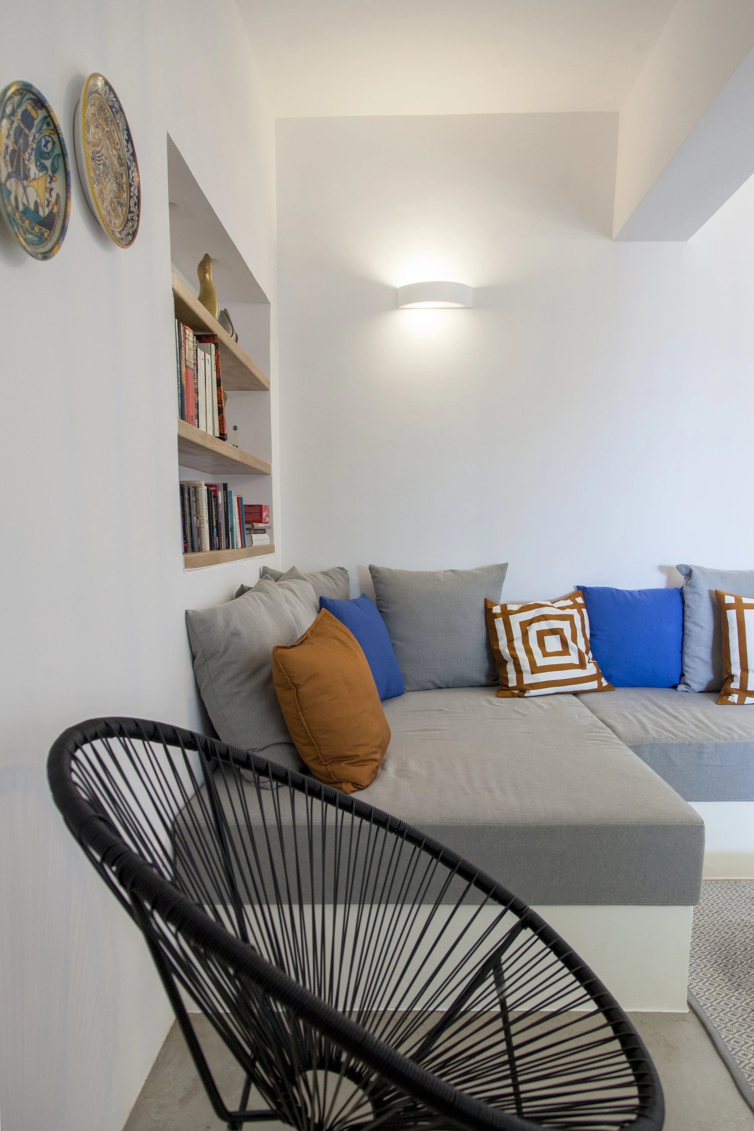Living room with a light grey sofa decorated with orange and blue pillows and a small round patterned rug. A black wire armchair is in the foreground. A built-in bookshelf on the wall has books and decorative items, with three decorative plates hangi