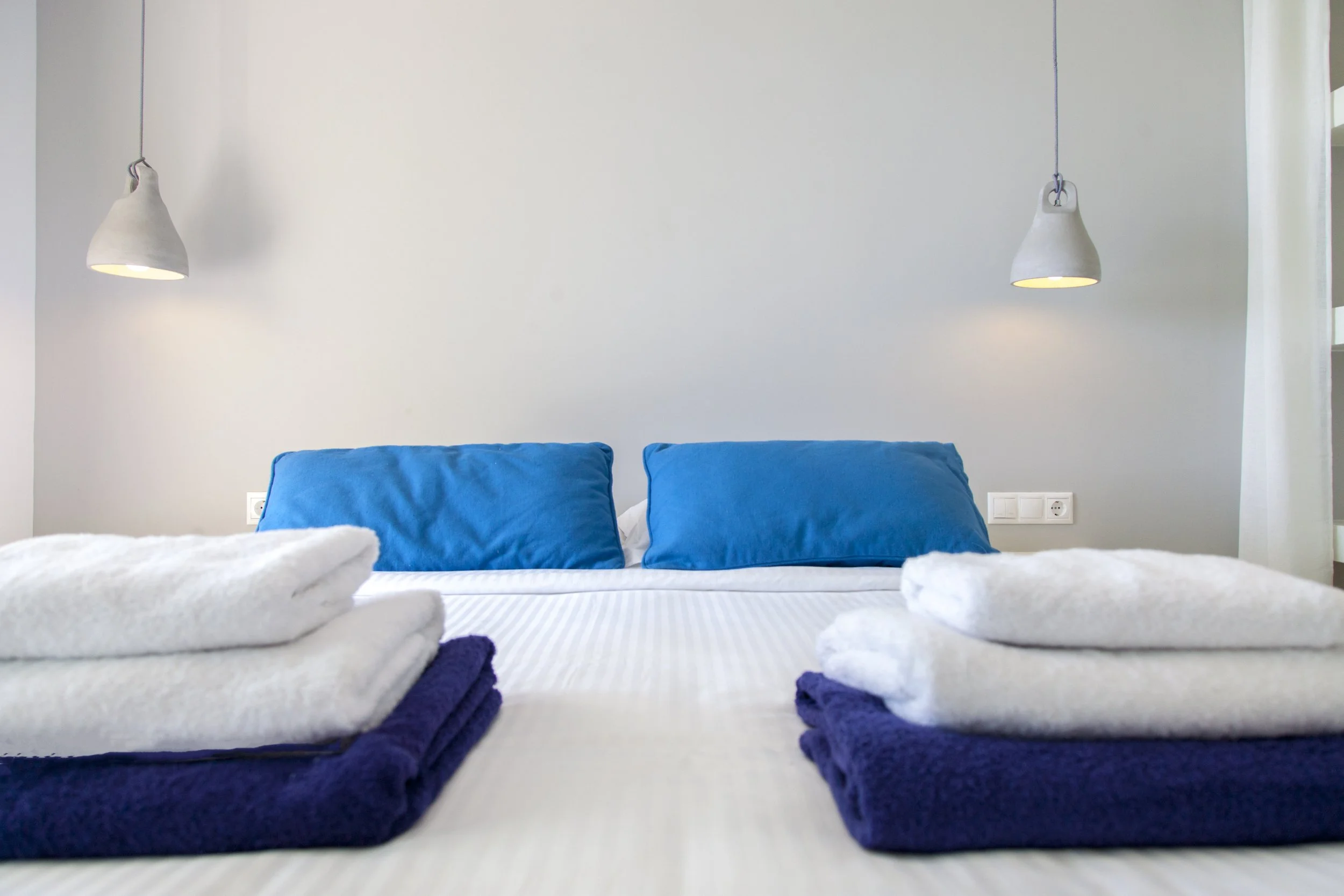 A neatly made bed with two blue pillows, white bedding, and two folded dark blue towels, in front of a white wall with two hanging white pendant lights.