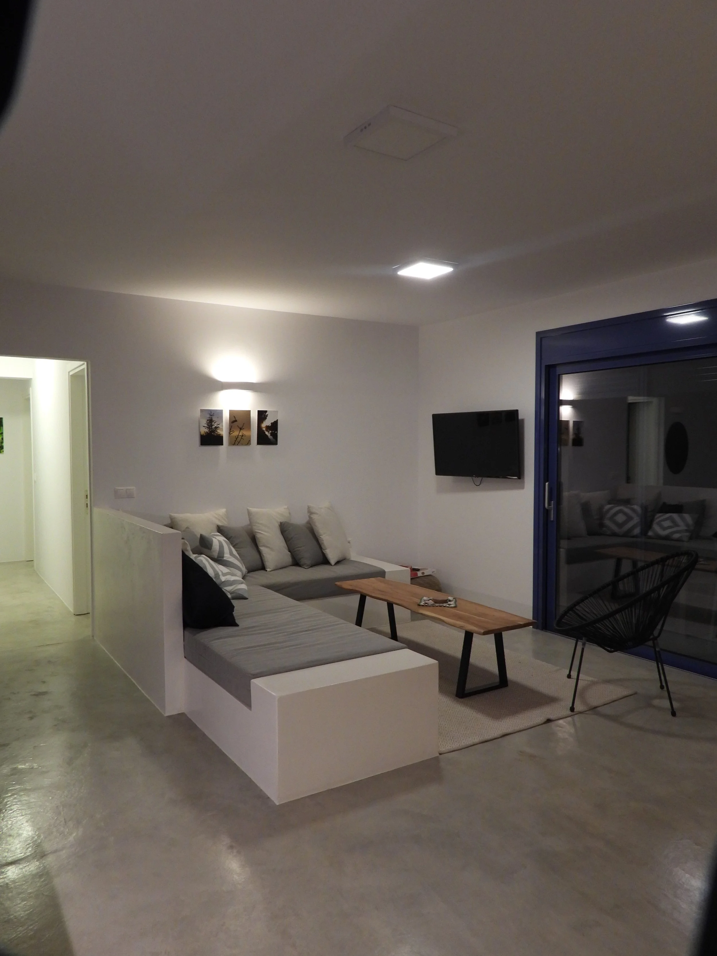 Living room with a gray sectional sofa with several pillows, a wooden coffee table, a black wire chair, a wall-mounted TV, a rug, and a dark window; wall art featuring tree silhouettes and a window with a blue frame.
