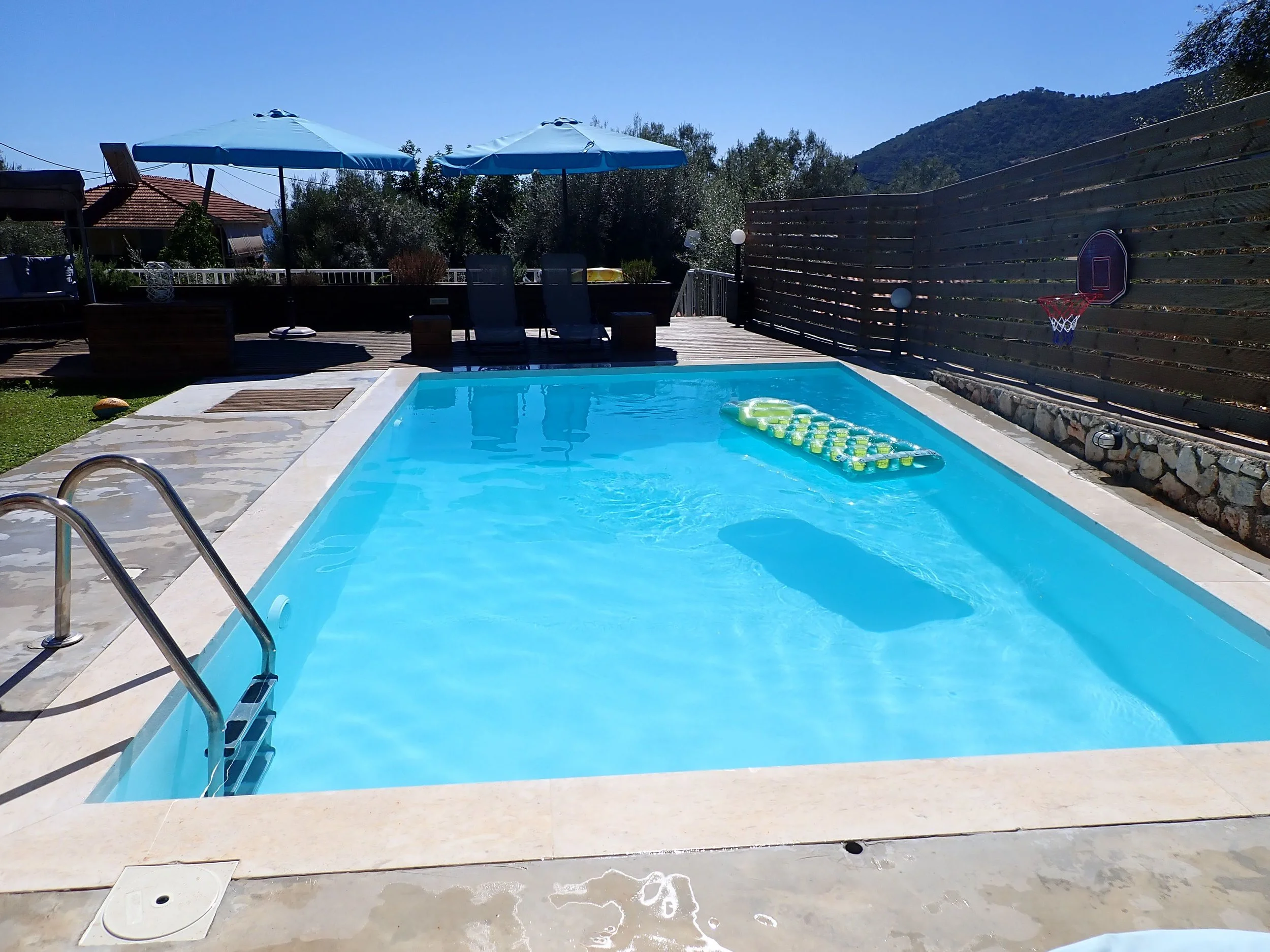A backyard swimming pool with blue water, surrounded by a stone deck and a wooden fence. There are two lounge chairs, two large blue umbrellas, and a small basketball hoop attached to the fence. In the background, there are hills and trees under a cl