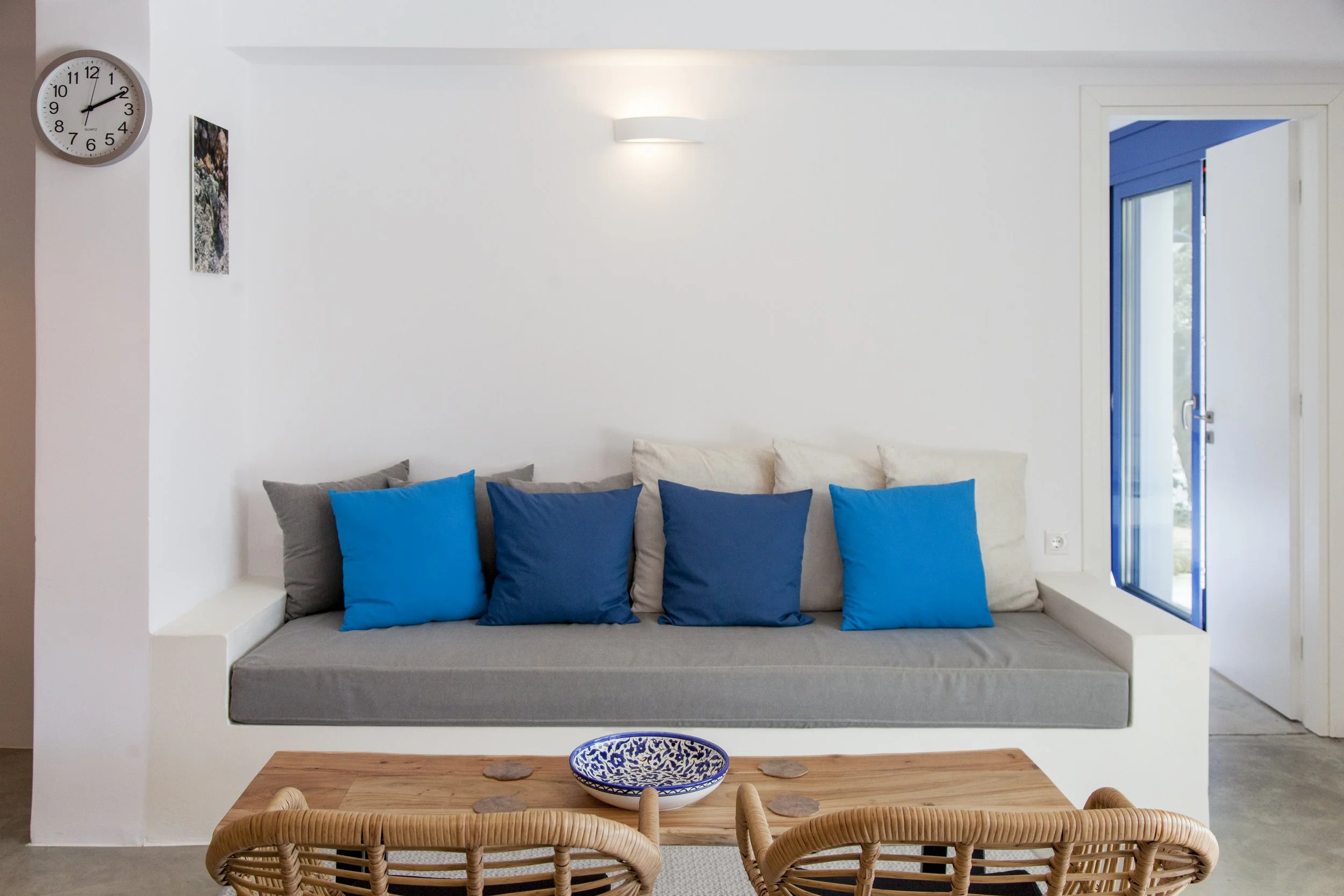 A living room corner with white walls, a white wall-mounted light, a clock on the left, a small painting, a window with blue shutters, and a comfortable gray sofa with blue and gray pillows. A wooden dining table with woven chairs and a decorative bl