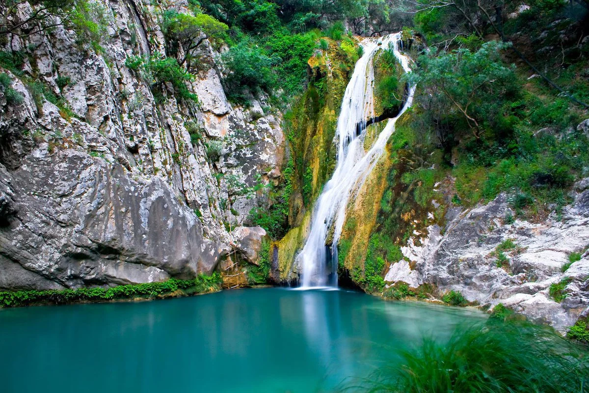 A scenic waterfall flowing down moss-covered rocks into a turquoise pool surrounded by lush green vegetation.