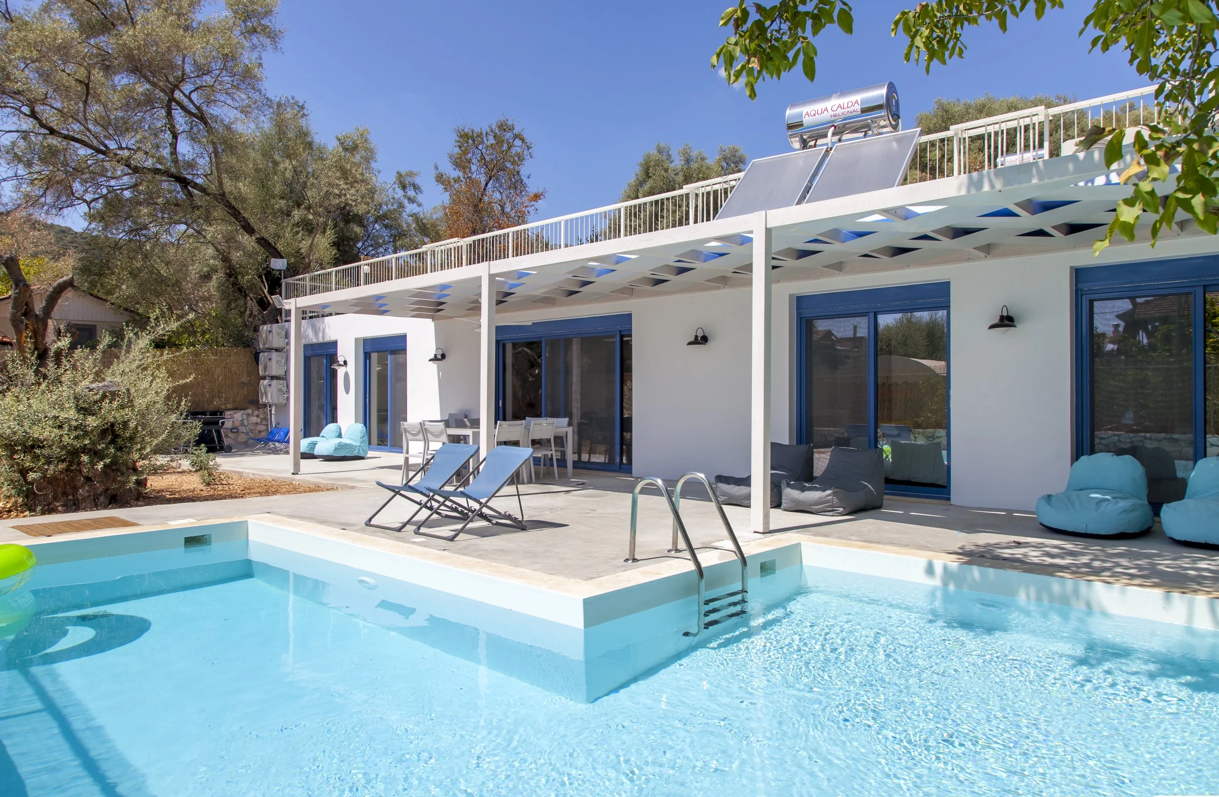 A backyard with a swimming pool, lounge chairs, bean bag chairs, and a dining table with chairs, adjacent to a blue and white house with large glass sliding doors. There are trees and bushes around the yard, with a clear blue sky overhead.