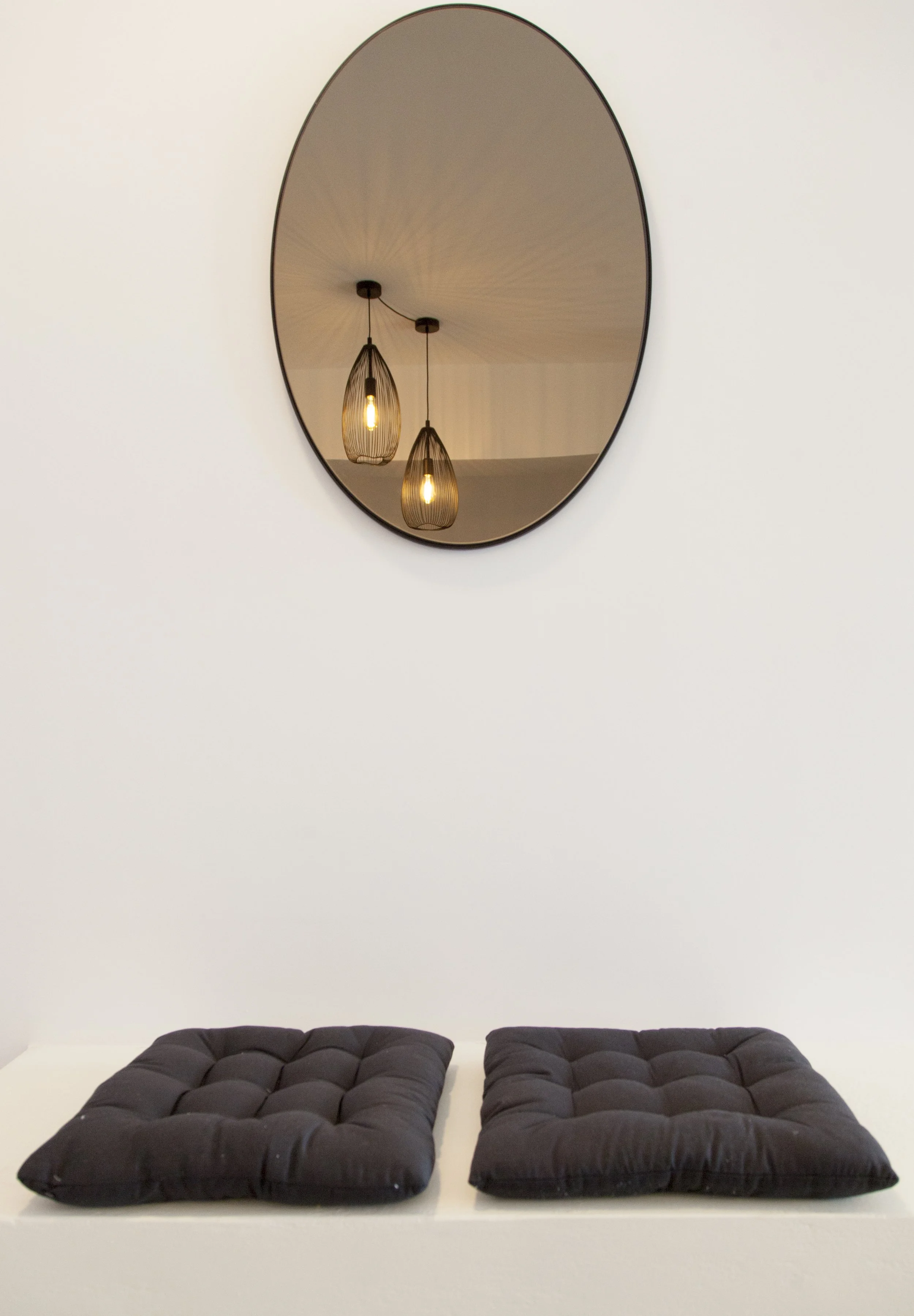 Two black cushions with tufted tops on a white surface in front of a white wall, with a large oval mirror and two pendant lights reflected in the mirror.