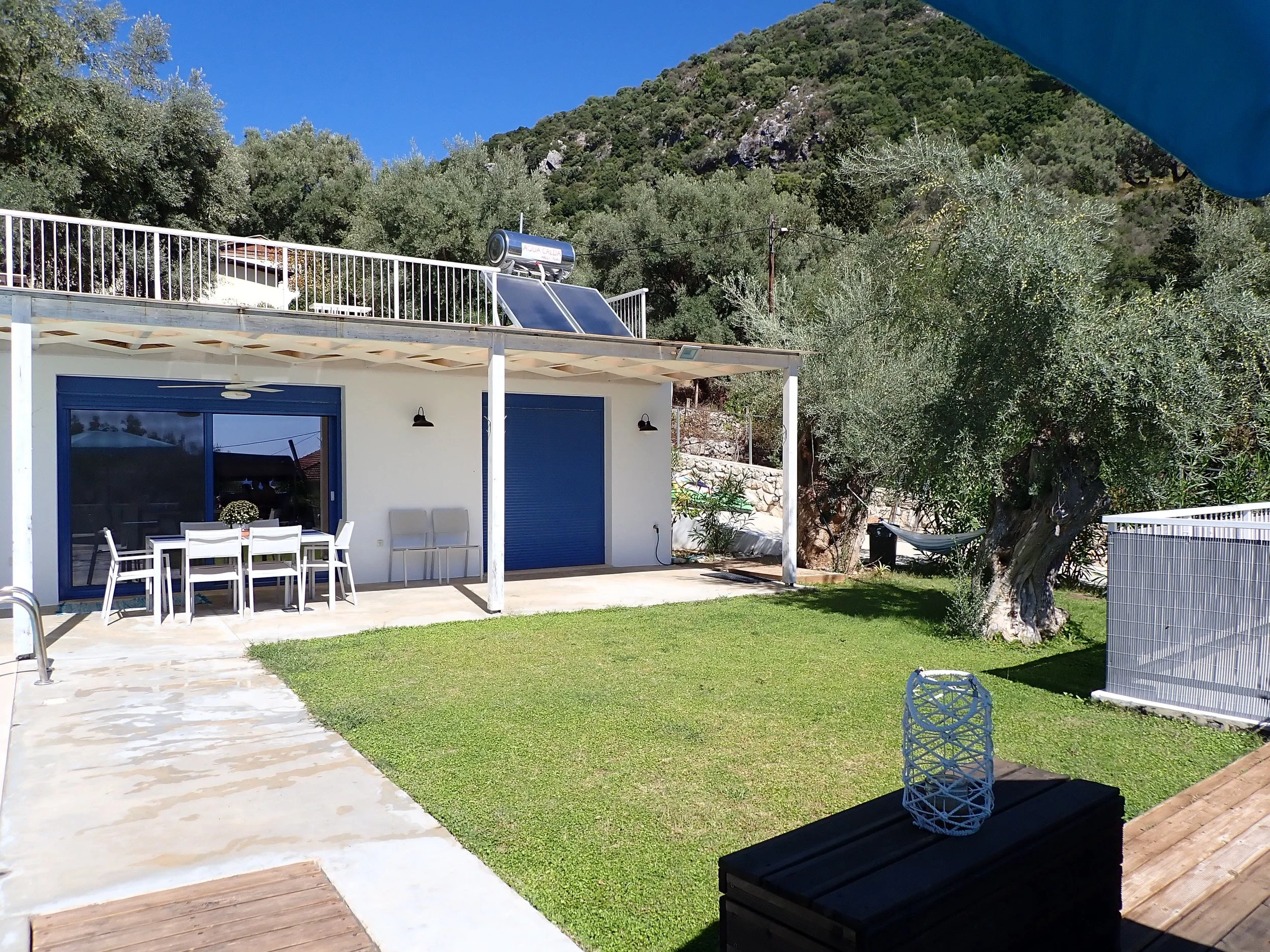 A backyard patio with a dining table and chairs, a swing, a manicured lawn, and a house with blue doors and windows. A mountain and trees are in the background under a clear blue sky.