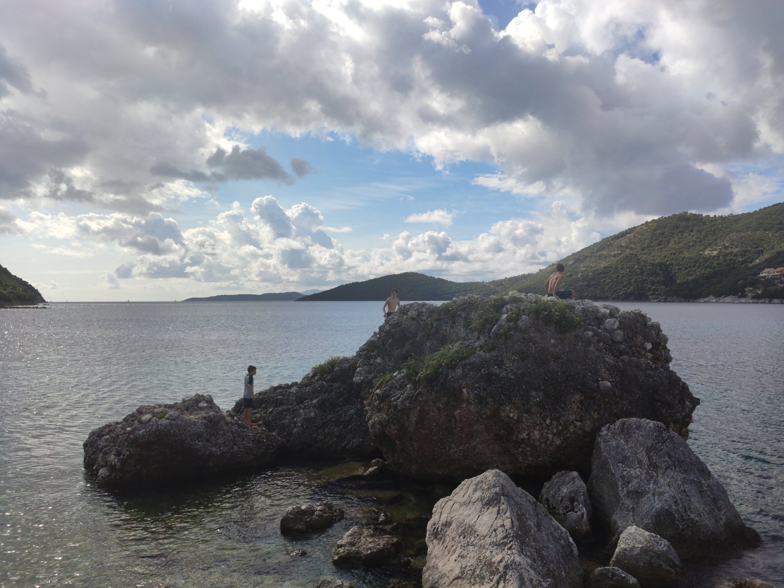 People swimming and standing on rocks by a lake with hilly landscape and cloudy sky.