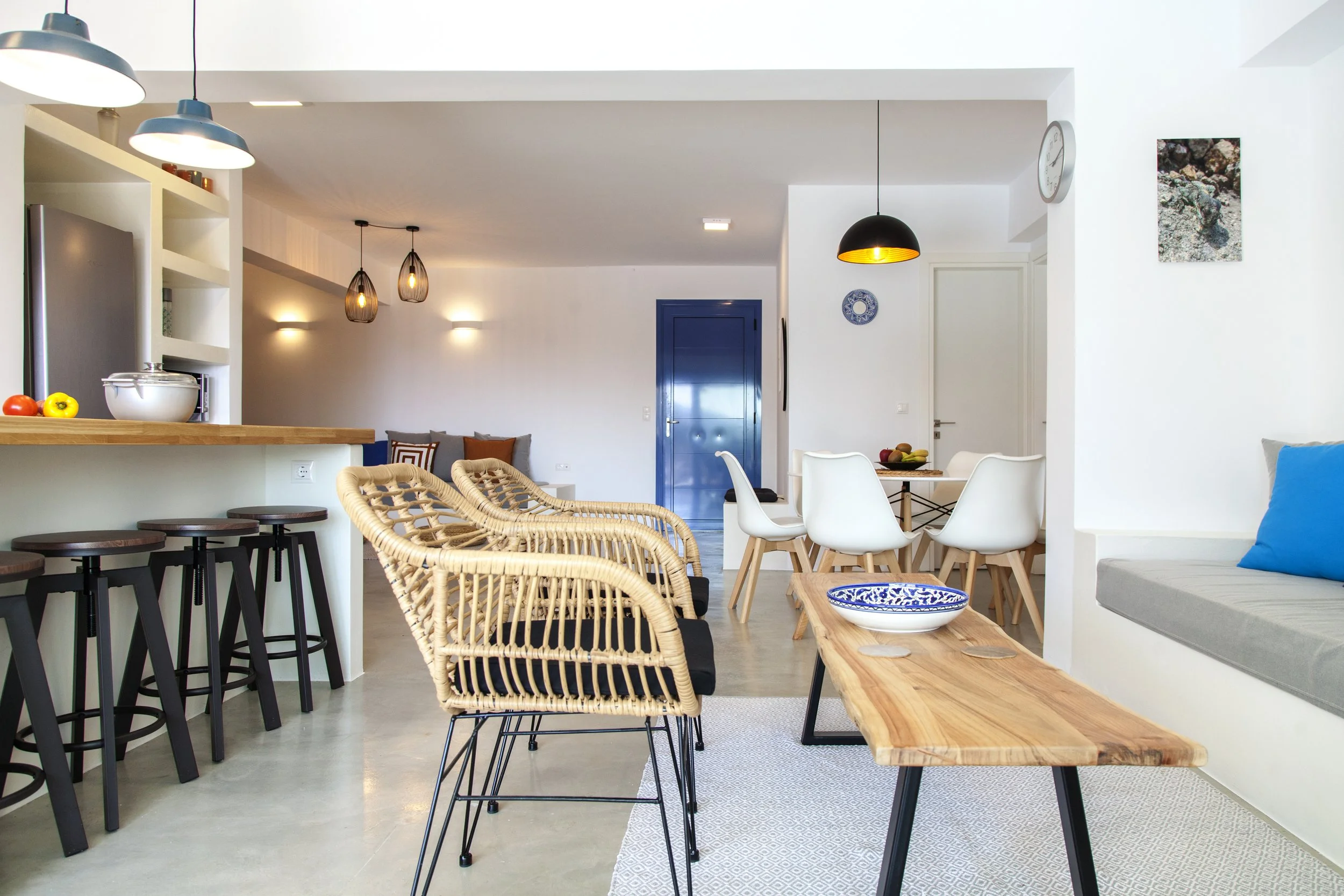 Modern open-concept living and dining area with white walls, blue accents, wicker and wooden furniture, pendant lighting, and decorative wall art.